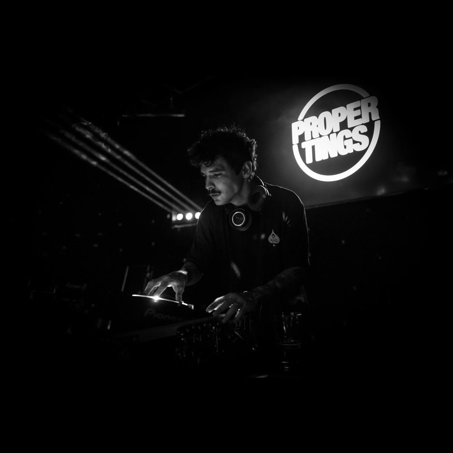A DJ performing at a club with a 'Proper Tings' sign illuminated on the wall behind him.
