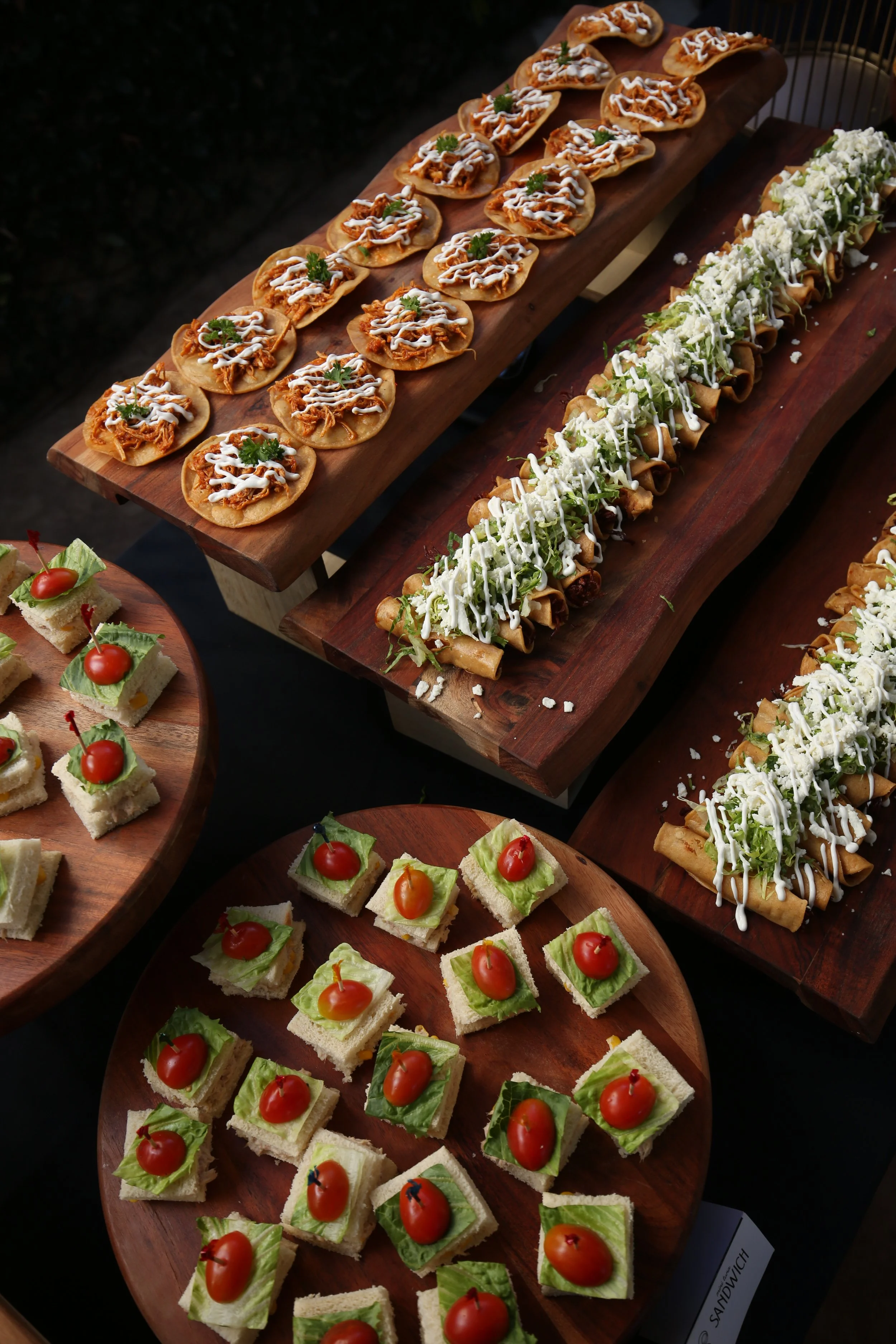 Assorted appetizers on wooden platters, including mini sandwiches with cherry tomatoes, lettuce, and shrink wrap, and topped tortilla chips with sauce and herbs.