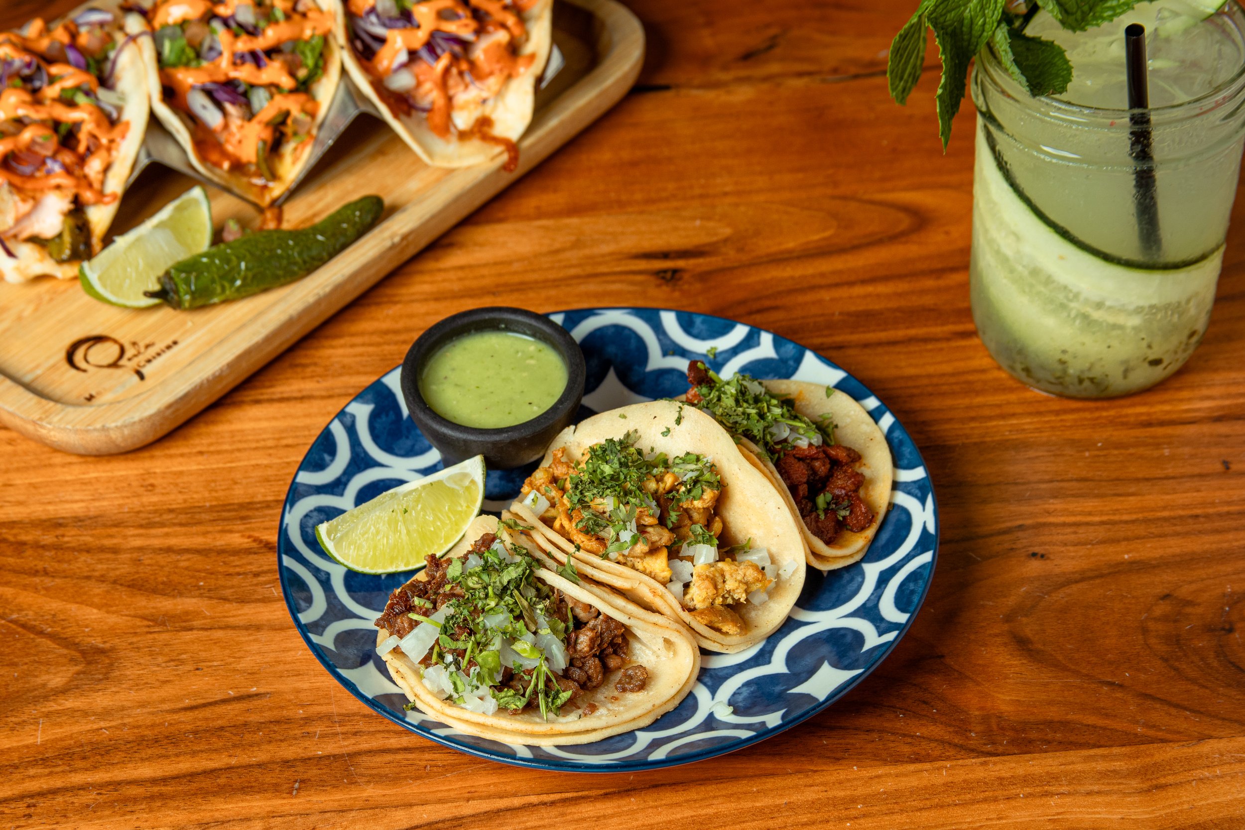 A plate of three tacos with chopped onions and cilantro, accompanied by lime wedges and a small bowl of green salsa on a wooden table. In the background, there is a tray of tacos and a cucumber-lime drink garnished with mint.