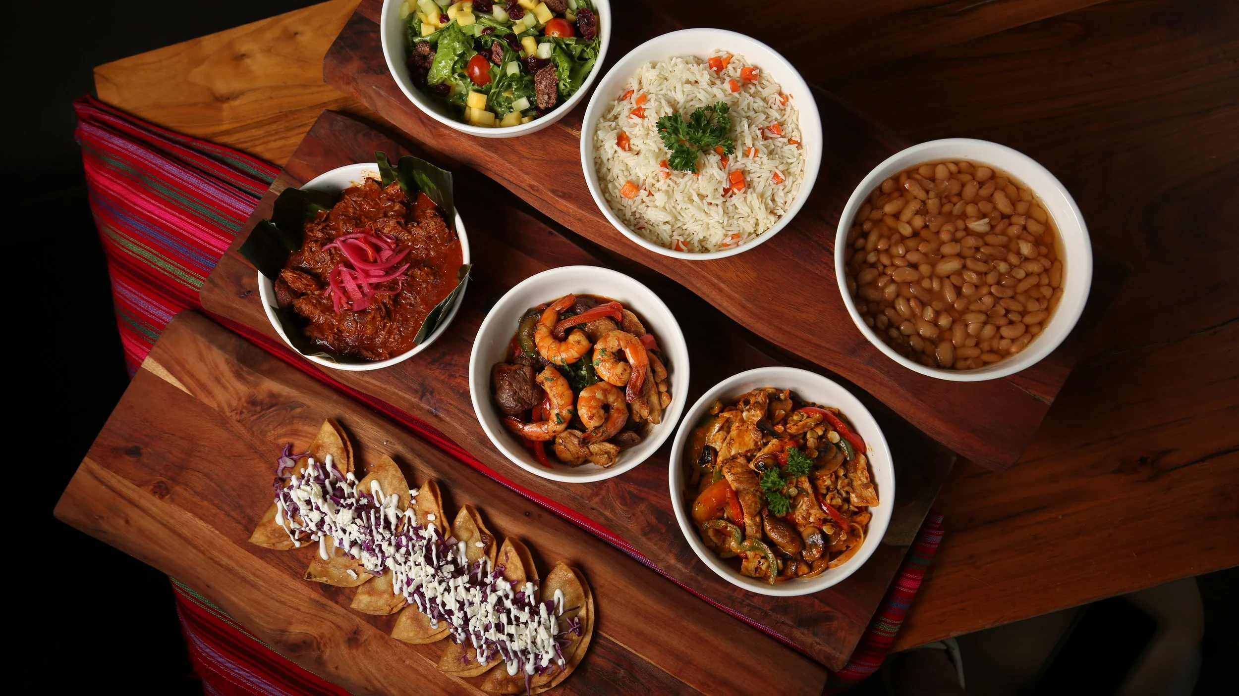 A wooden serving board with a variety of Mexican dishes, including bowls of rice, beans, and various stews, and a plate of nachos with dipping sauces, all arranged on a table with a striped cloth.