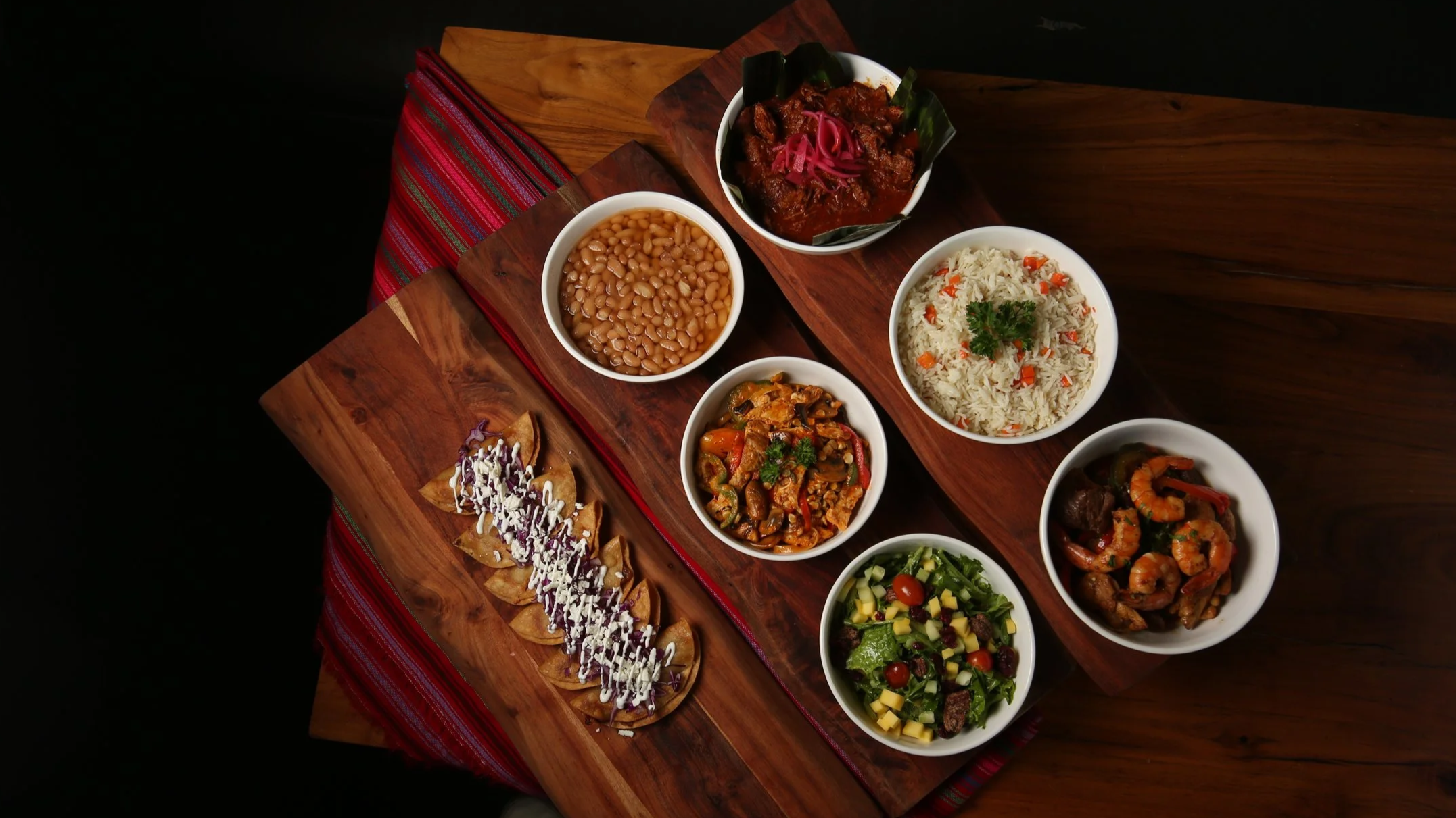 Assorted Mexican dishes on a wooden table, including tacos with toppings, rice, beans, beef stew, shrimp, and vegetable salad.