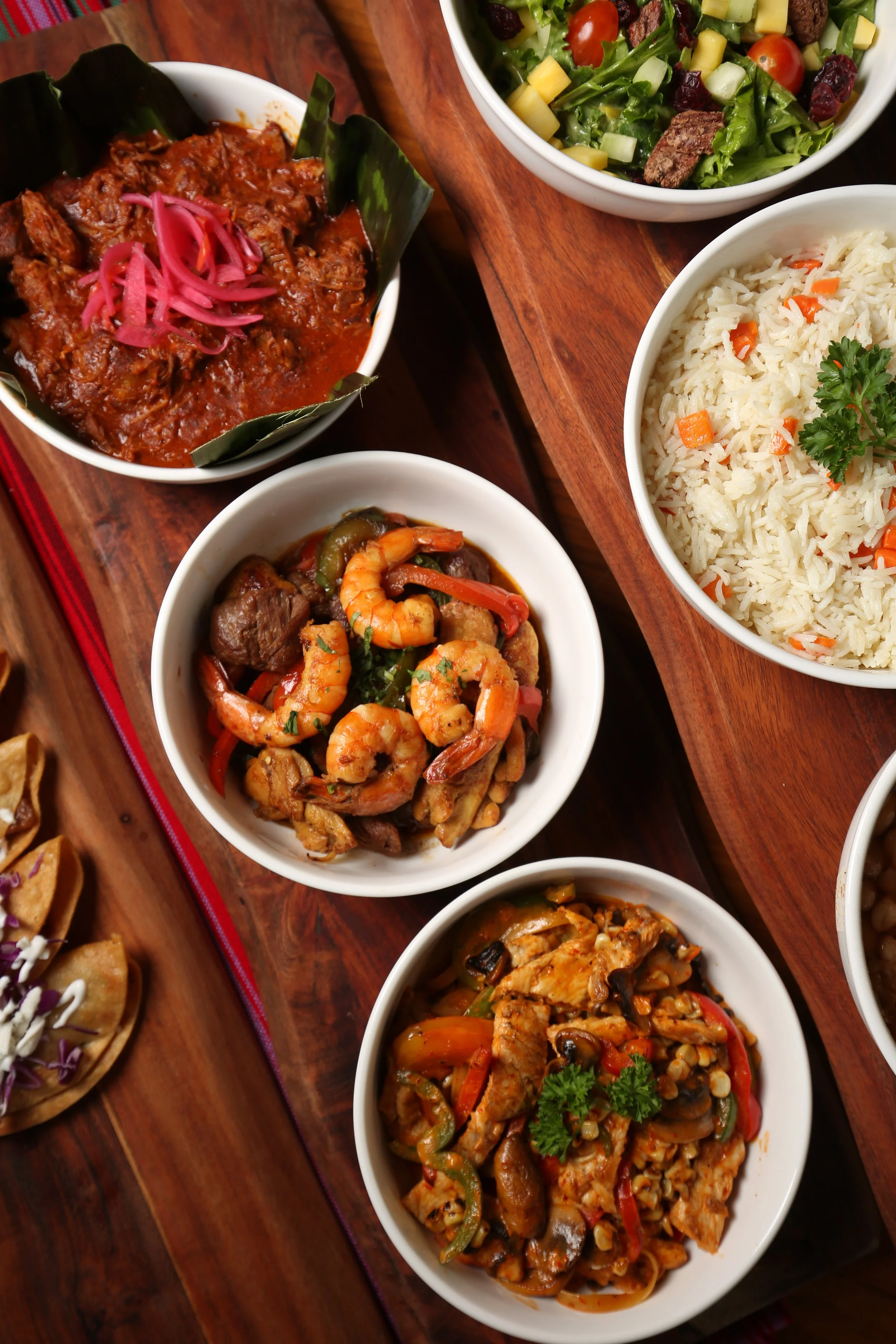 A variety of small bowls of Mexican dishes on a wooden table, including shrimp with vegetables, shredded chicken in sauce with cilantro, beef with chili peppers, and beef with vegetables, along with bowls of rice and salad.