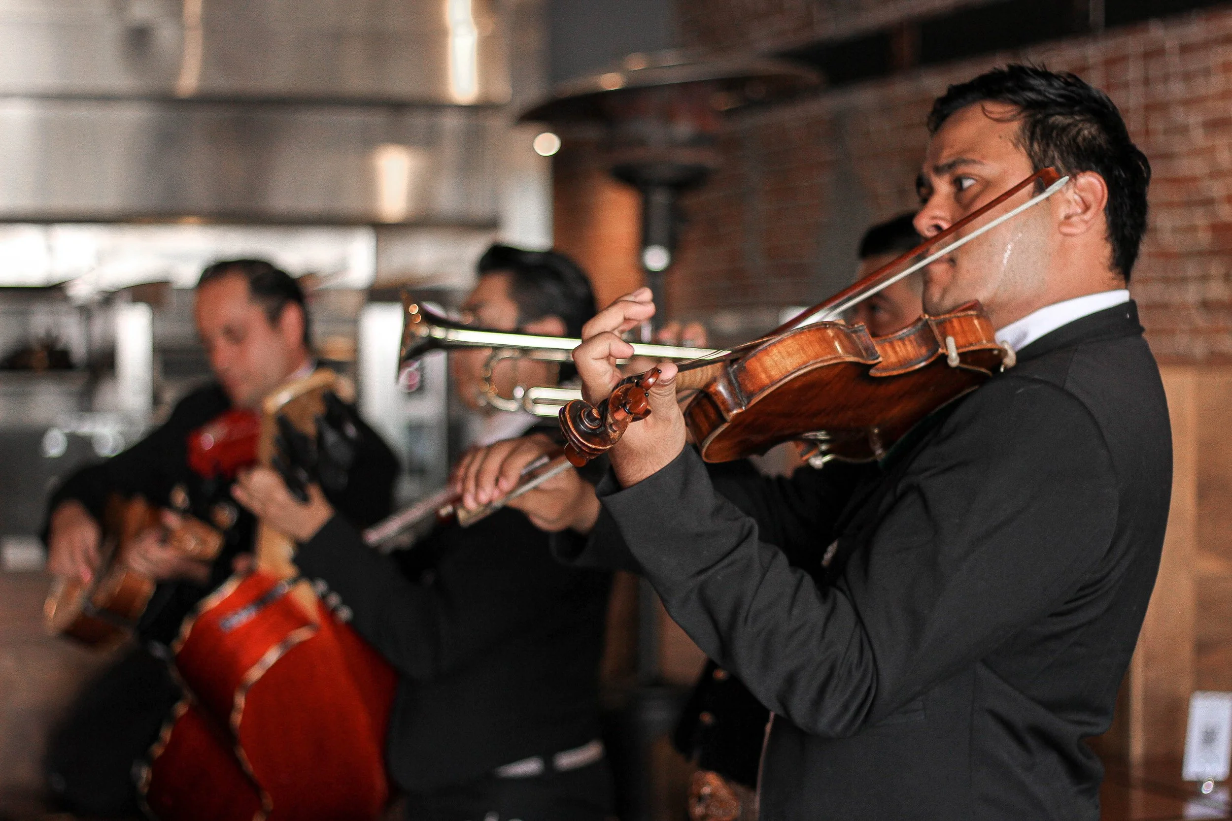 Group of musicians playing string instruments, including violins and a drum, in a restaurant with brick walls.