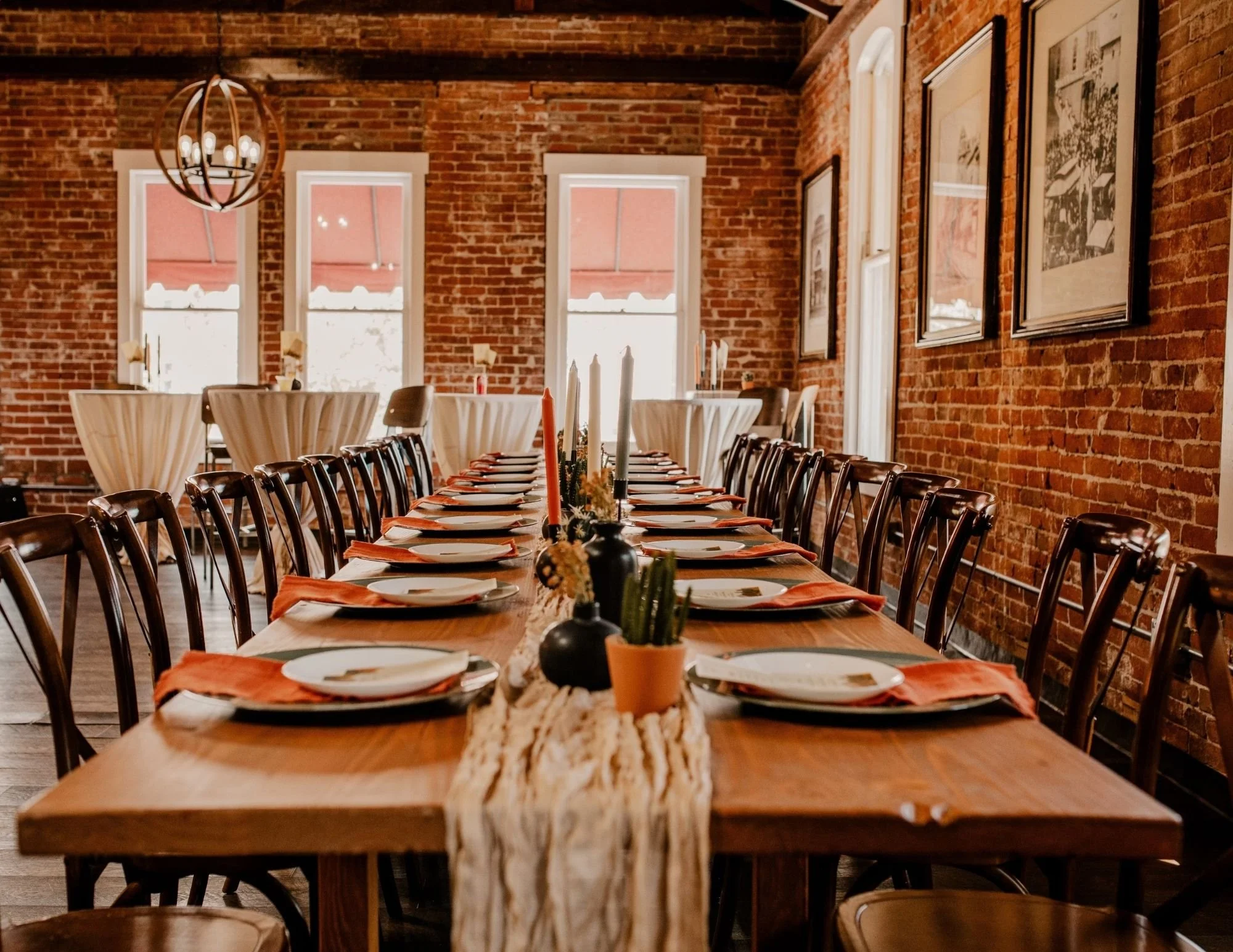 A long wooden dining table set for a meal with white plates, orange napkins, and black candle holders with candles. There are potted plants and candelabras on the table. The room has exposed brick walls, large windows, framed artwork, and modern lighting fixtures.