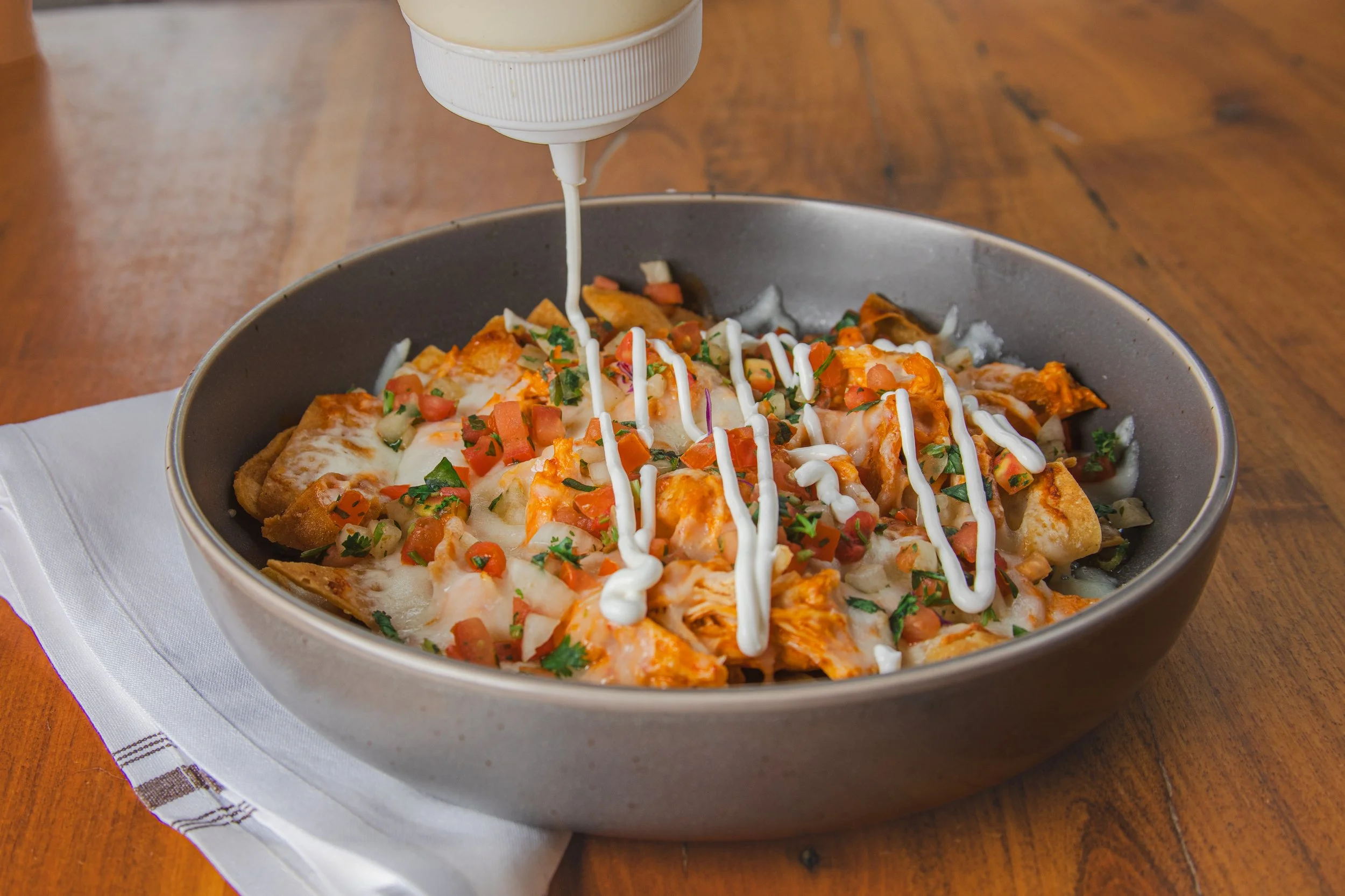 A bowl of nachos topped with cheese, diced tomatoes, chopped herbs, and drizzled with sour cream being poured from a squeeze bottle, set on a white napkin on a wooden table.