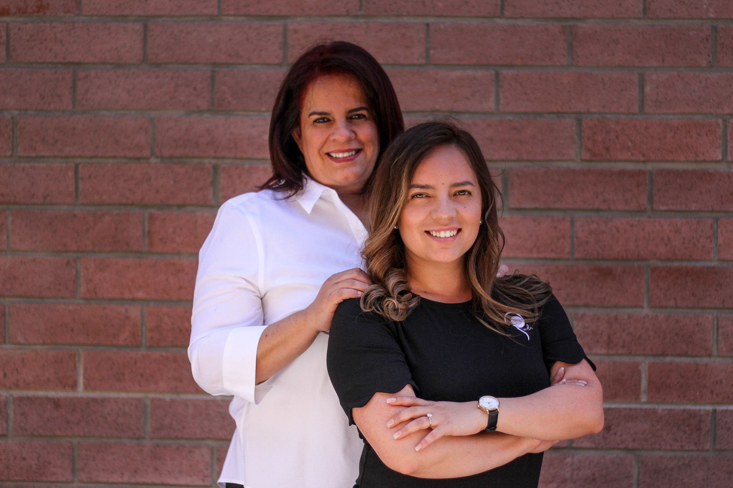 Two women standing in front of a brick wall, smiling at the camera. One woman has shoulder-length dark brown hair and wears a white shirt. The other woman has dark blonde hair with curls and wears a black top, a watch on her left wrist, and an engagement ring on her left hand.