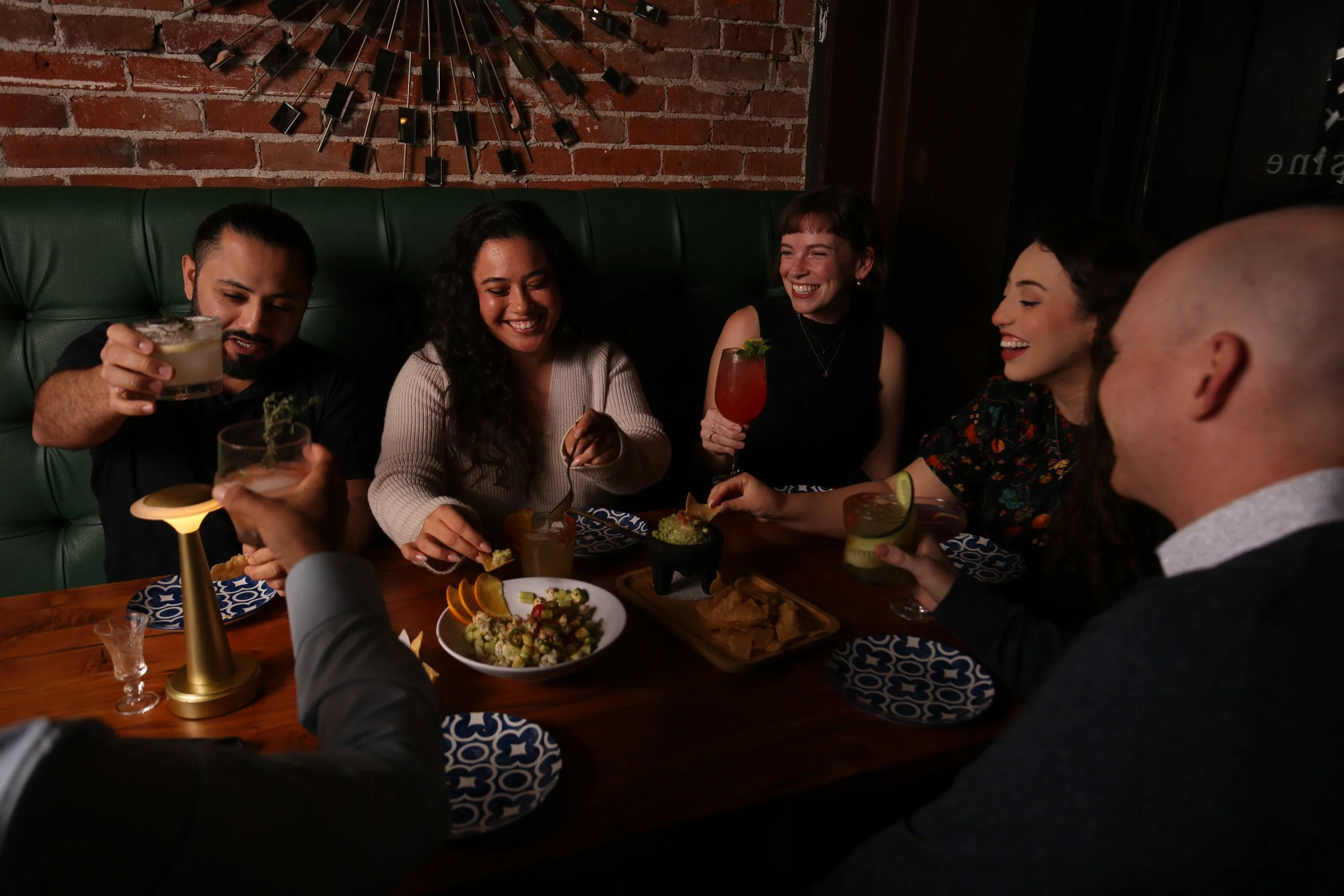 A group of six friends enjoying drinks and food together at a restaurant table, laughing and socializing.