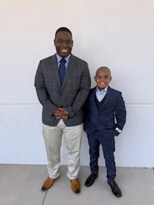 Two males standing against a white wall, one adult and one child, both dressed in suits and smiling.