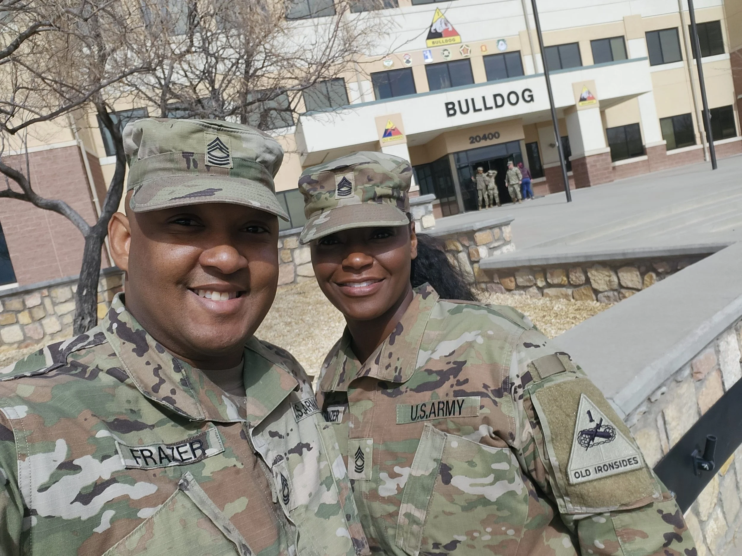 Two U.S. Army soldiers in camouflage uniforms and hats smiling in front of a building with a sign that reads 'BULLDOG'.