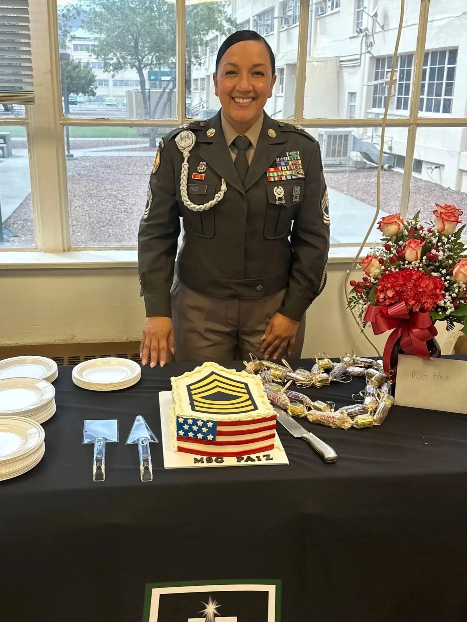 A woman in a military uniform standing behind a table with a decorated cake, plates, and a bouquet of flowers, celebrating an event.