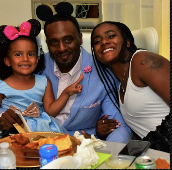 A family of three sitting in a restaurant, smiling at the camera. The young girl on the left wears a blue dress and Minnie Mouse ears headband. The man in the middle wears a light blue blazer, a pink shirt, and Minnie Mouse ears headband. The woman o