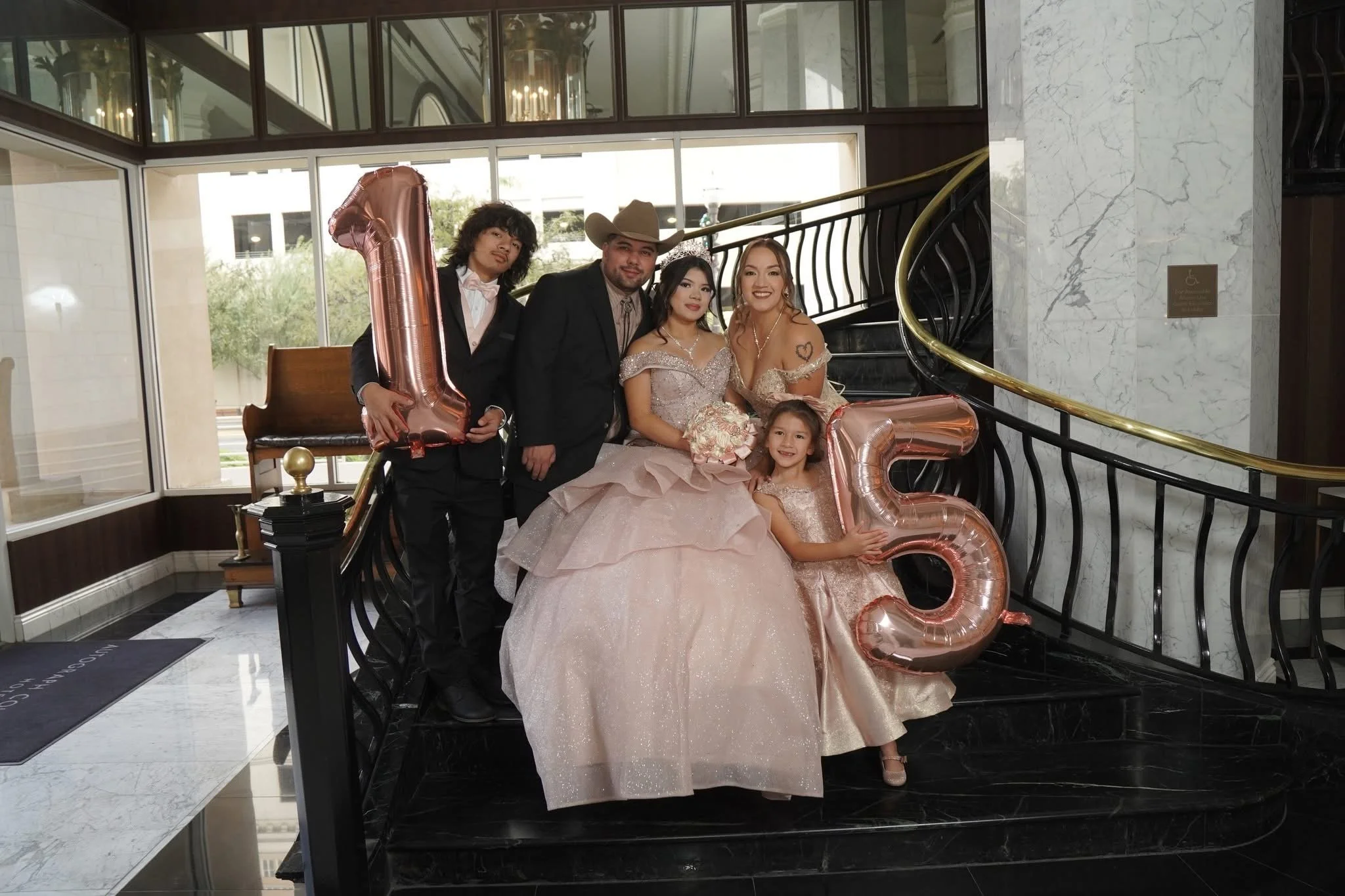 Group of five people celebrating at a wedding reception, with some holding large rose gold balloons spelling out 15, standing on a black marble staircase in front of a glass door with a chandelier visible inside.