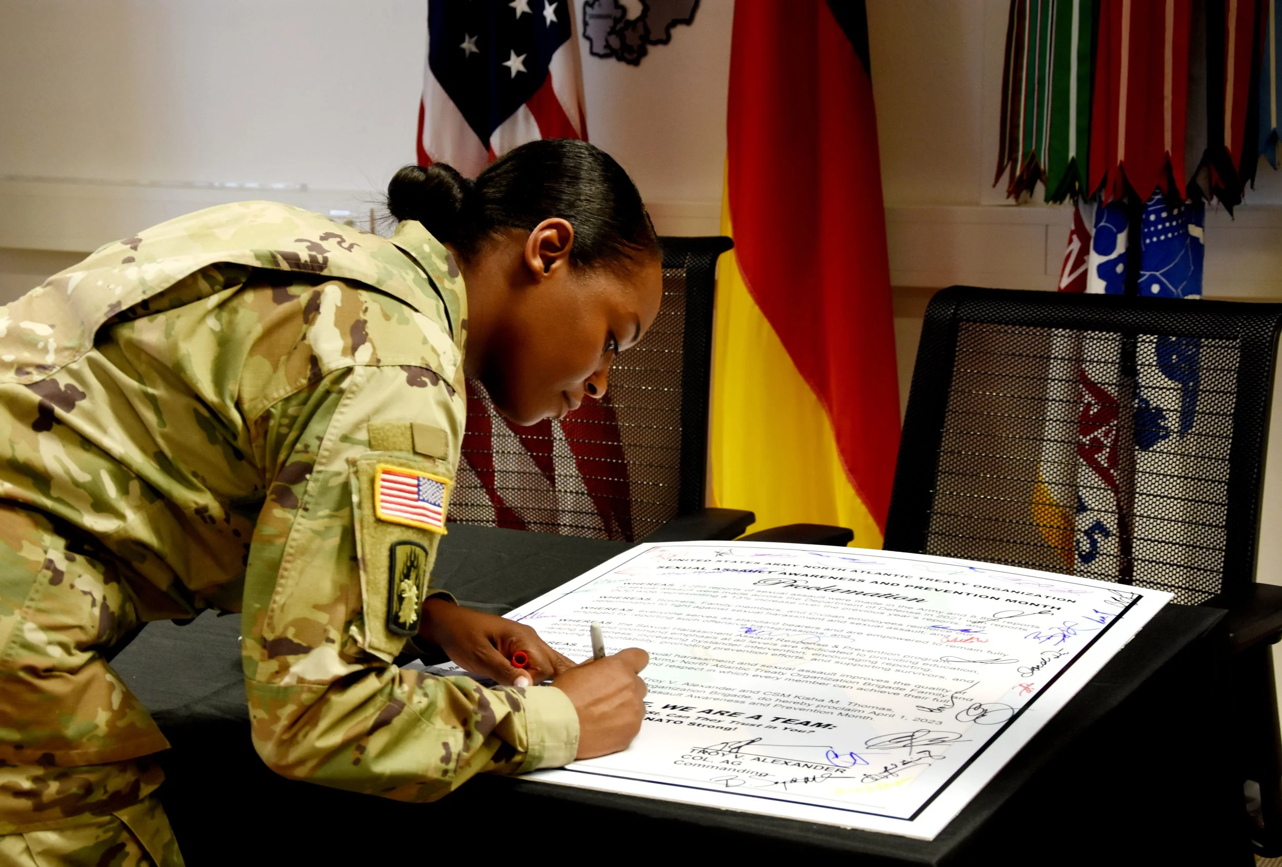 A woman in military uniform signing a document on a table, with flags in the background.