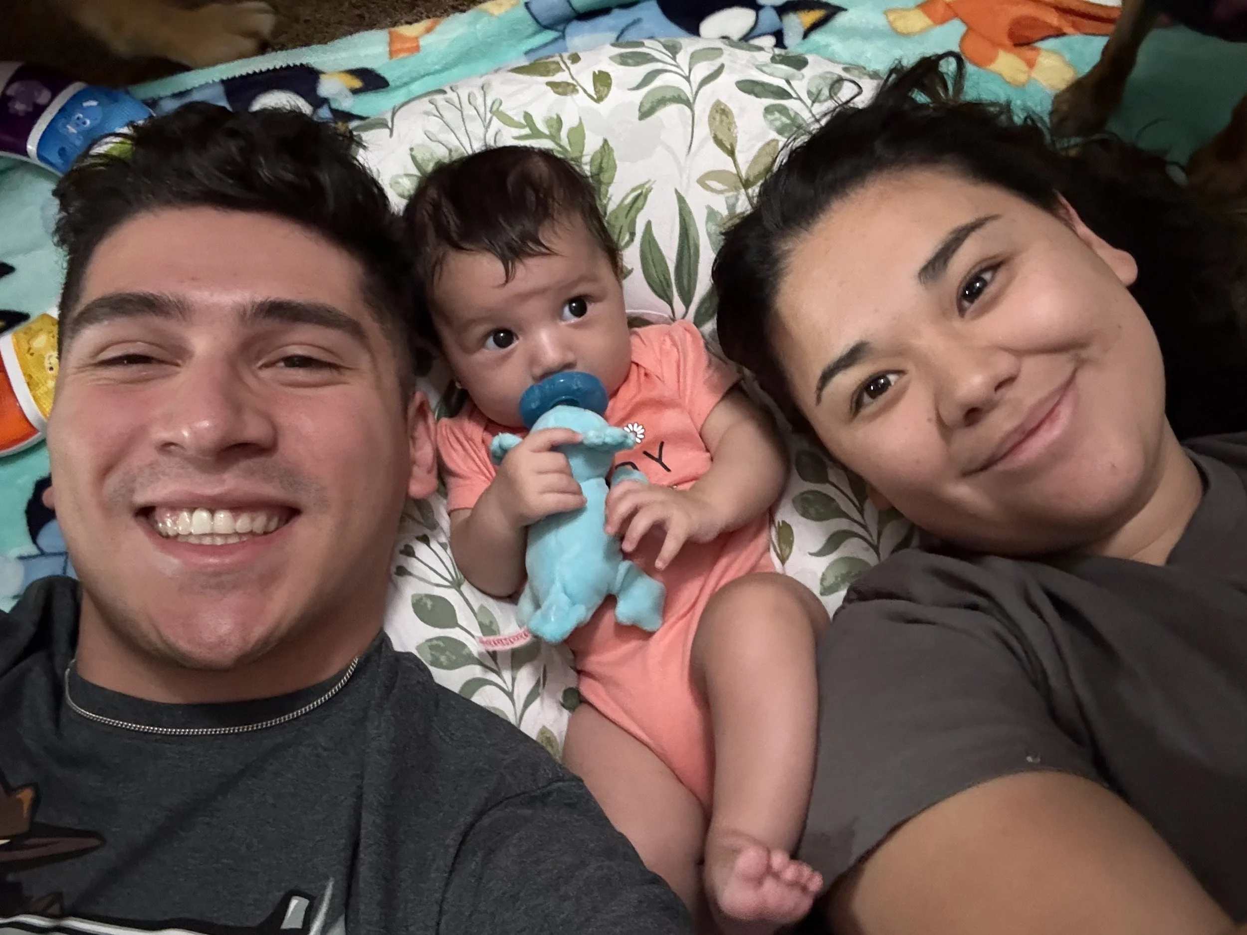 A smiling young man with short dark hair, a young woman with long dark hair, and a baby girl lying on a floral-patterned pillow. The baby has a blue pacifier and is holding a blue plush toy. All are taking a selfie together, appearing happy and relax