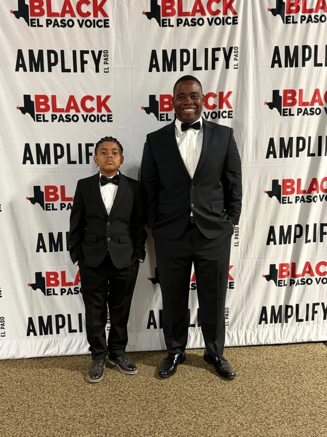 Two males standing against a white wall, one adult and one child, both dressed in suits and smiling.