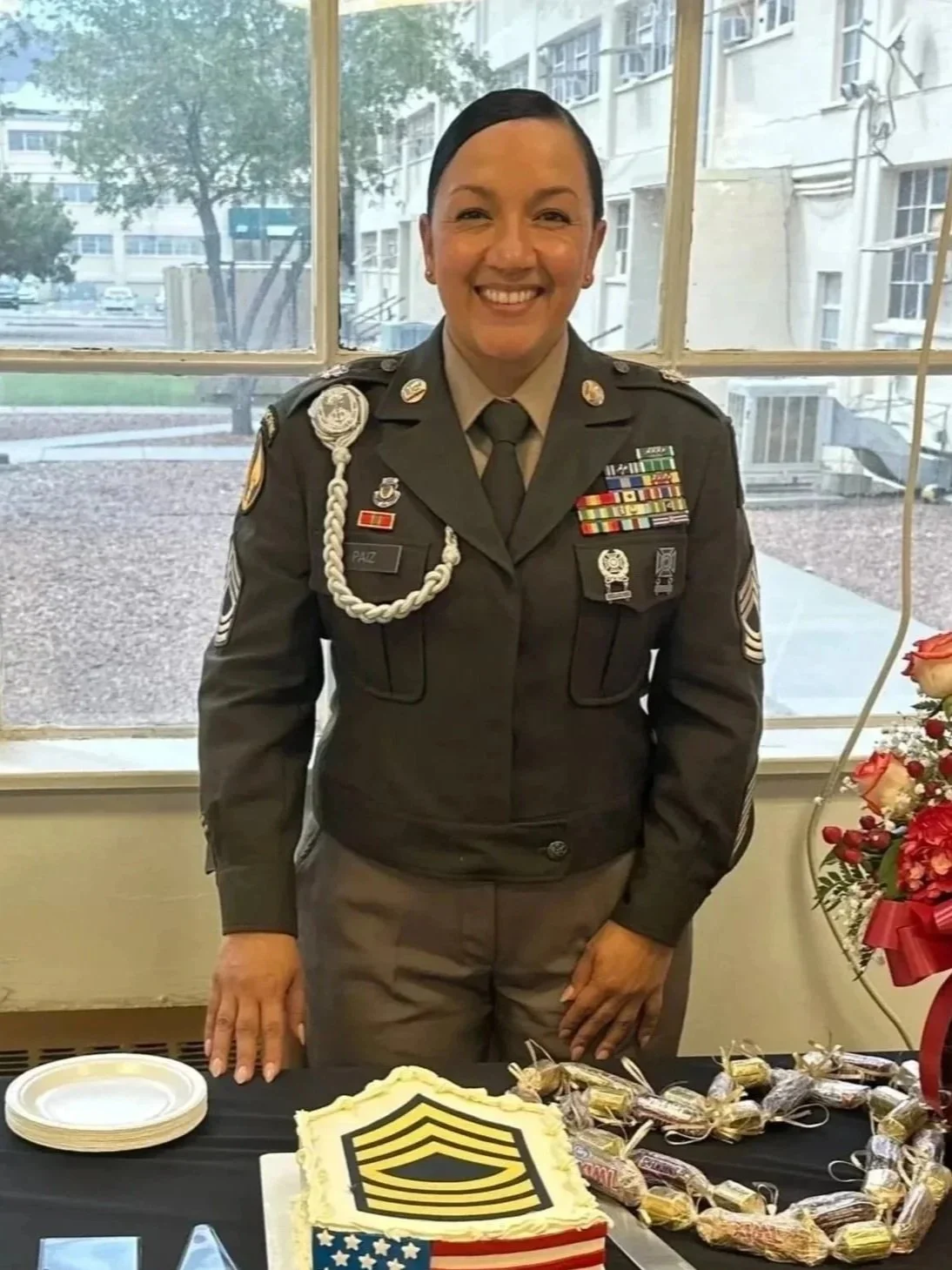 A woman in a military uniform standing behind a table with a decorated cake, plates, and a bouquet of flowers, celebrating an event.