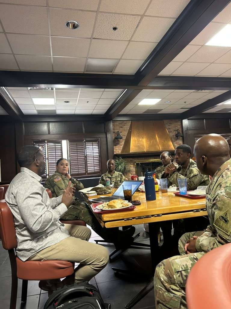 Group of six military personnel and one civilian woman seated around a dining table engaged in conversation in a restaurant with brick and wood interior and a brass fireplace hood.