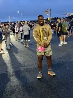Man standing with his hands clasped in front, smiling at an outdoor event during the evening, with a crowd of people and food trucks in the background.