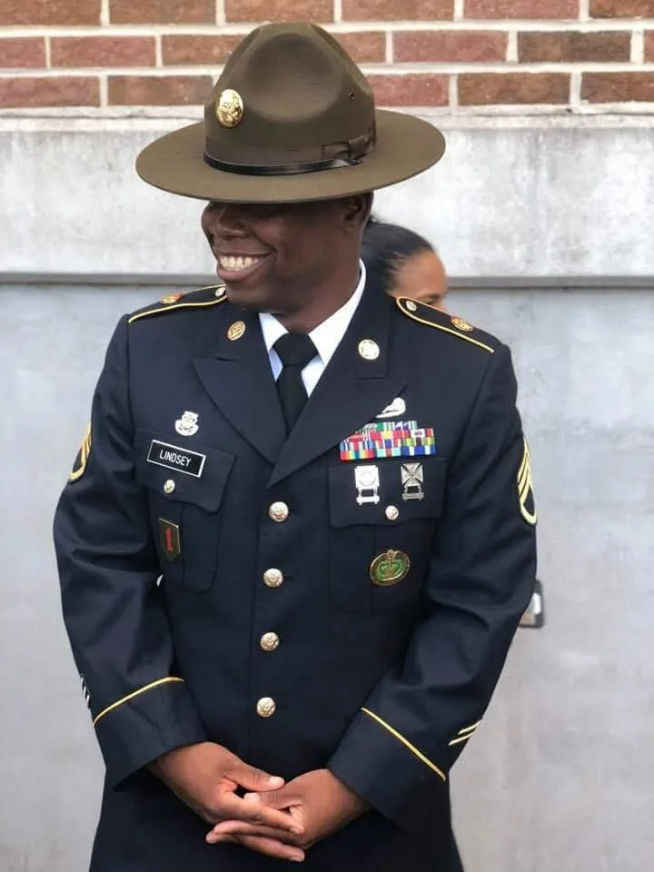 A smiling woman dressed in a U.S. Army dress uniform with medals, ribbons, and a nametag 'LINDSEY', wearing a drill sergeant hat with a badge, standing in front of a brick wall.