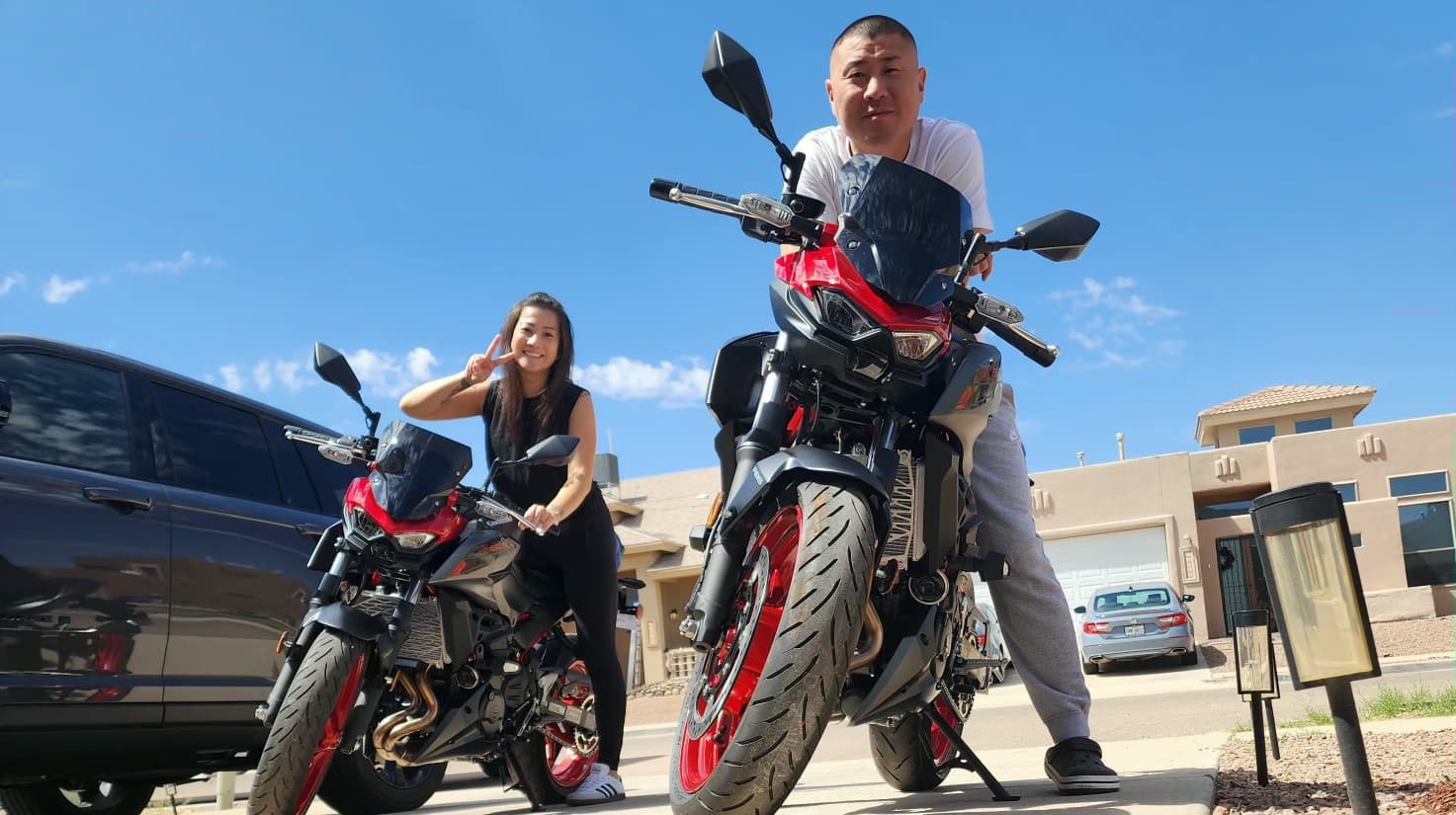 A man and a woman posing with motorcycles on a driveway on a sunny day. The man is leaning on his motorcycle, and the woman is smiling and making a peace sign.