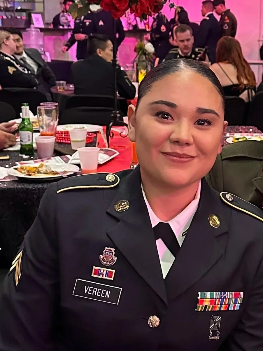 A young woman in a military uniform sitting at a banquet table with a smile, surrounded by people in similar uniforms at a formal event.