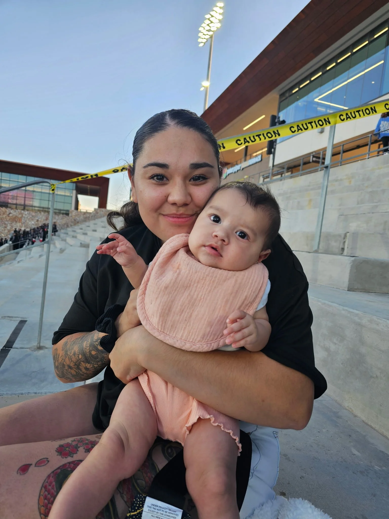 A young woman holding a baby at an outdoor stadium with caution tape and stadium lights in the background