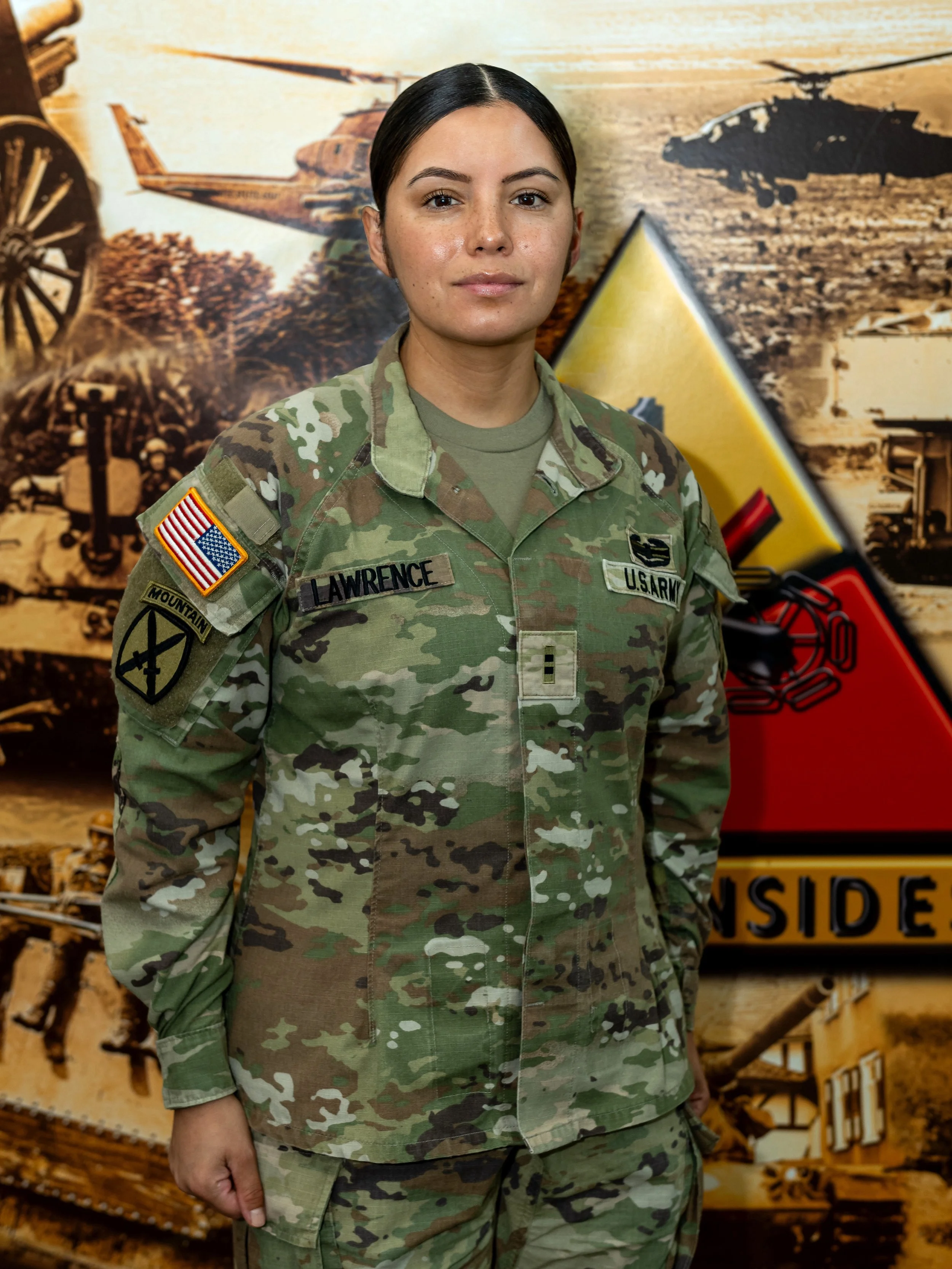 A woman in U.S. Army camouflage uniform standing in front of a military-themed backdrop with tanks and military vehicles.
