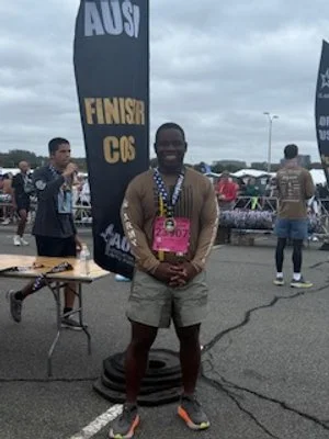 A man standing at a finish line event with a medal around his neck, wearing a pink race bib, in a parking lot with other people and flags in the background.