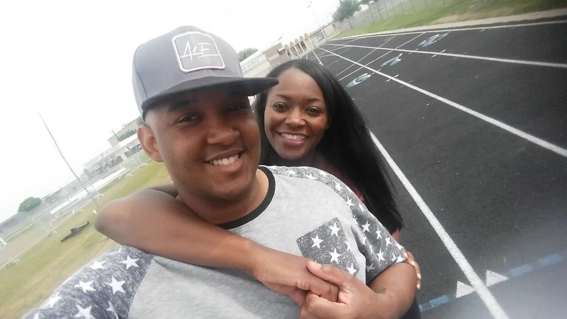 A happy couple taking a selfie on an outdoor running track with empty lanes and sports field in the background. The man is wearing a gray baseball cap and a gray t-shirt with star patterns, and the woman has long black hair and is smiling widely. The