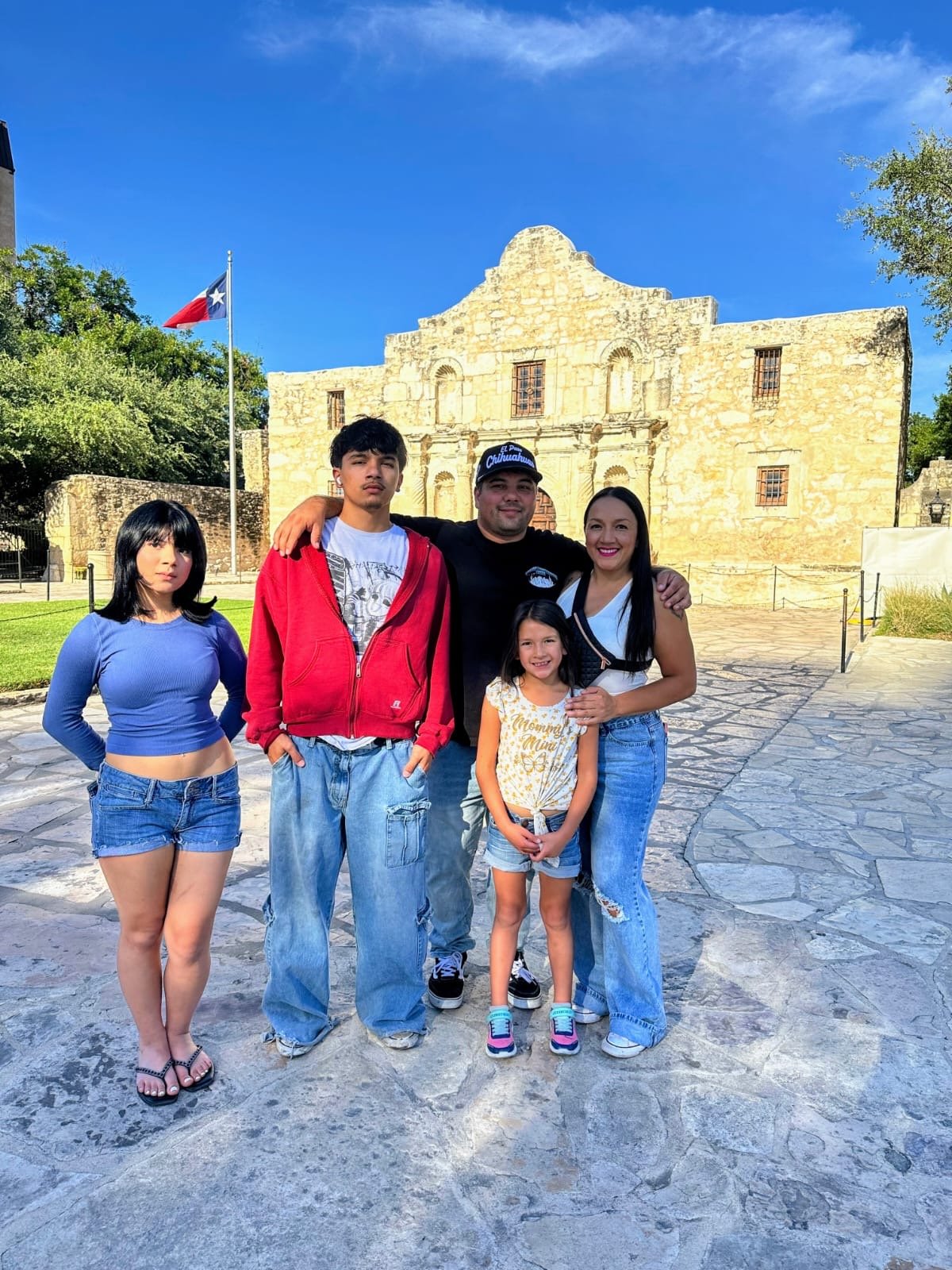 Family of five posing in front of the Alamo in San Antonio, Texas. The background features the historic stone building and a Texas flag is visible on the left. The sky is clear and blue.