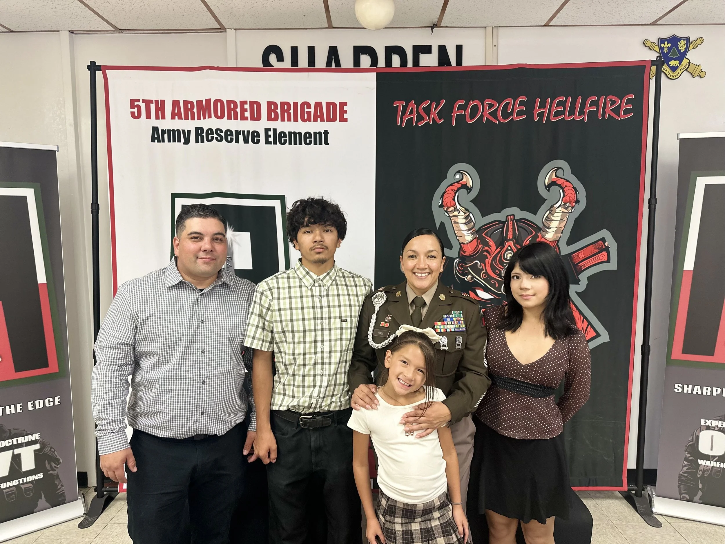 Group of five people, including a woman in military uniform, standing in front of a banner with military emblems and text about the 5th Armored Brigade, Army Reserve, and Task Force Hellfire, at an indoor event.