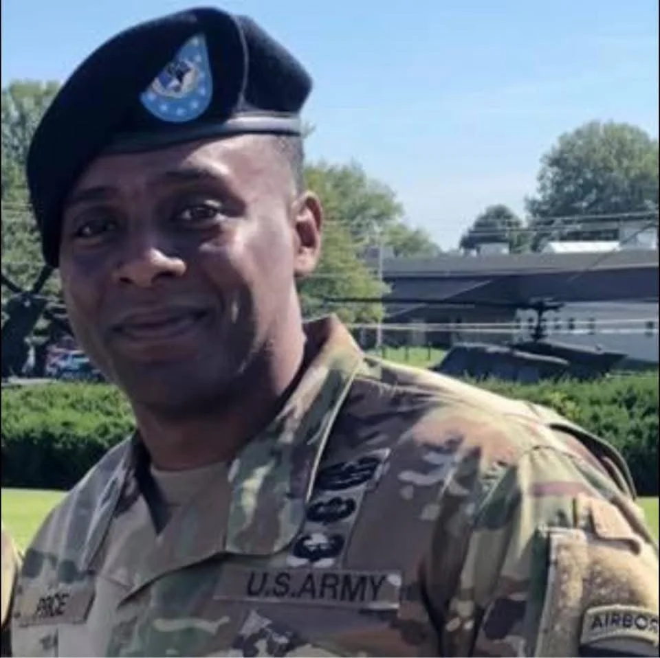 A smiling man in U.S. Army camouflage uniform and a beret standing outdoors with trees and buildings in the background.