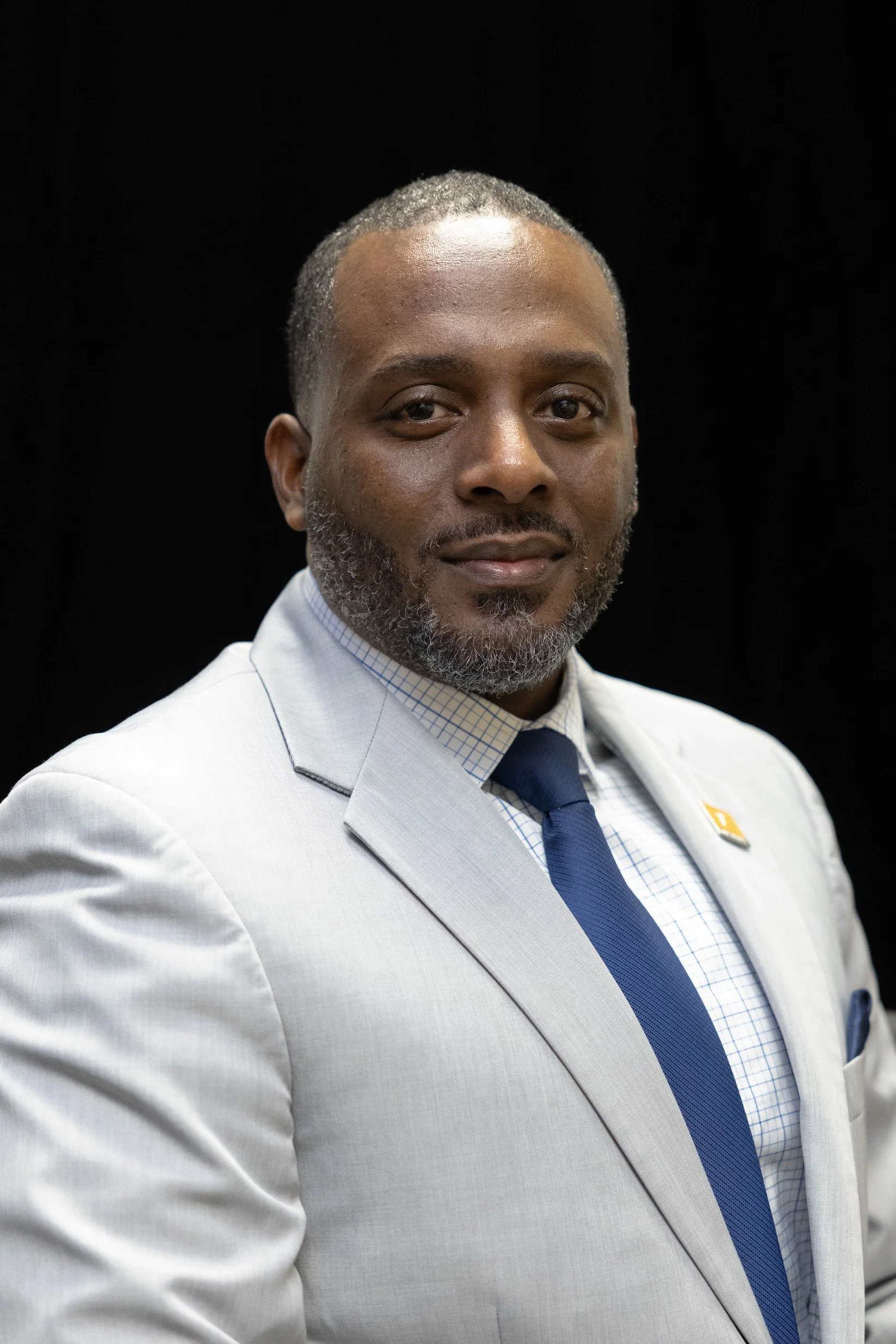A confident man in a light gray suit, checkered shirt, and dark blue tie, posing against a black background.