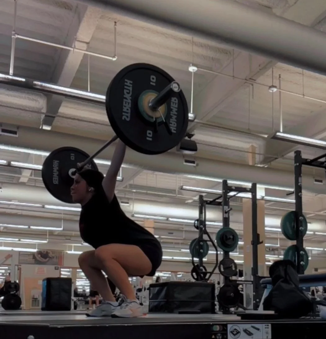 A woman in a gym performing a barbell squat, wearing black workout clothes, surrounded by gym equipment and mirrors.