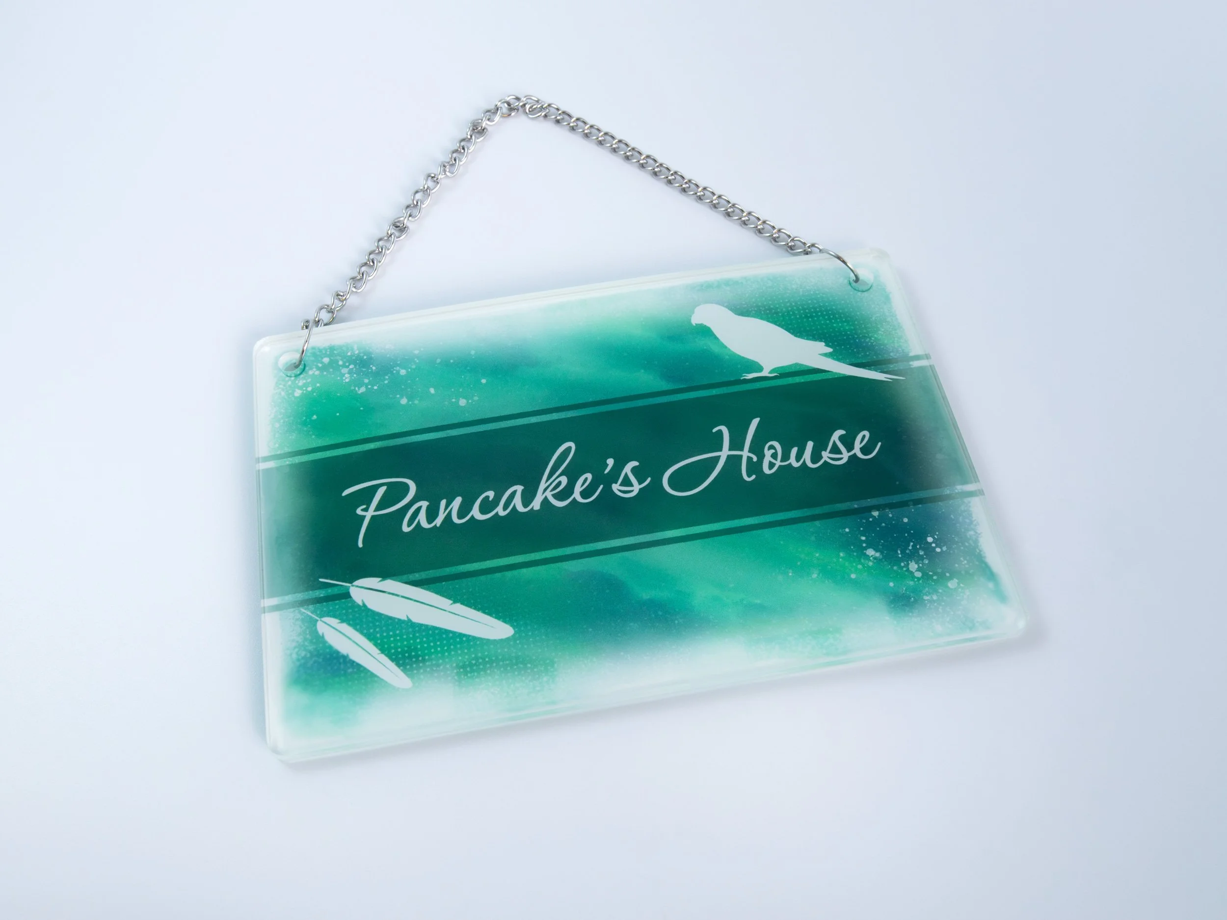 Top view of a small acrylic sign with a metal chain. It has bird-themed artwork with text that says "Pancake’s House"