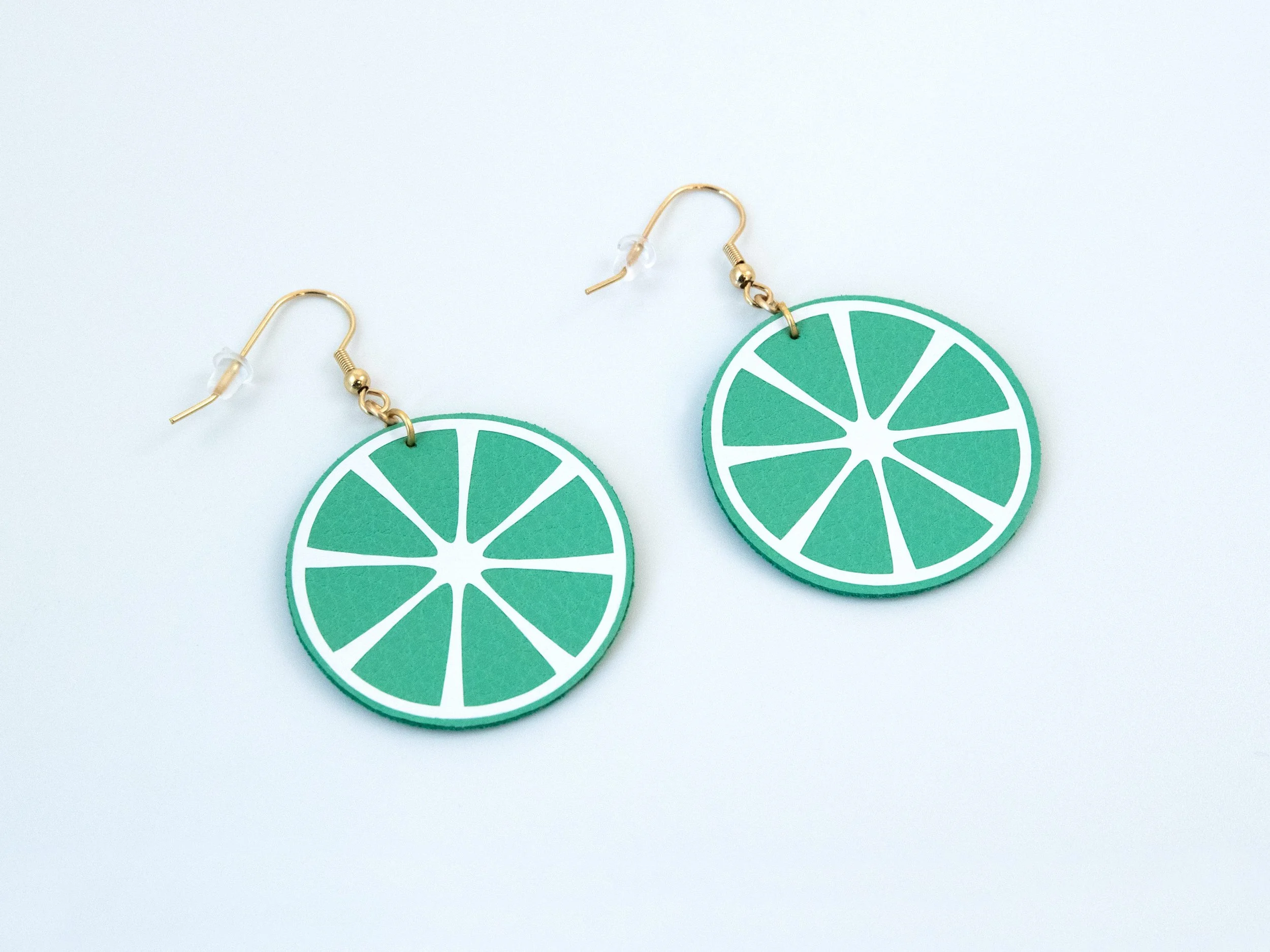 Angled view of round citrus slice earrings. They have green faux leather, white vinyl details, and gold-finished hooks