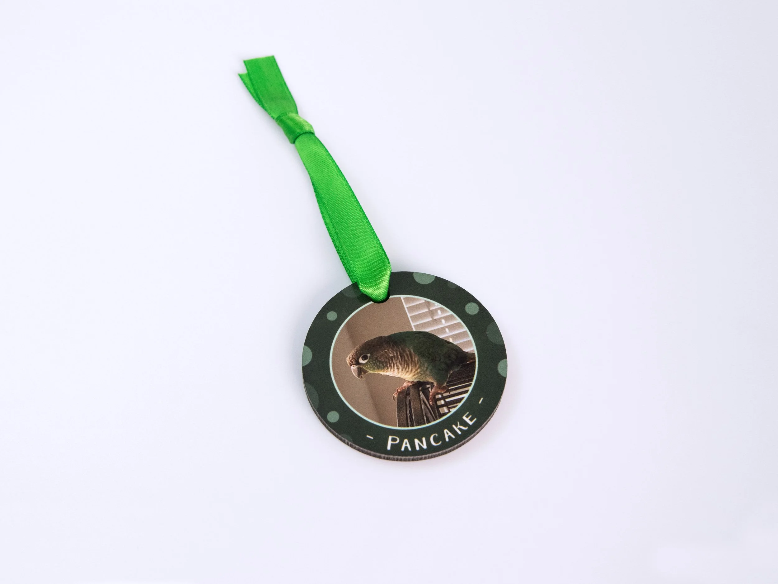 Top view of a sublimated MDF ornament with a ribbon. It has a conure photo, green color scheme, and the name "Pancake"