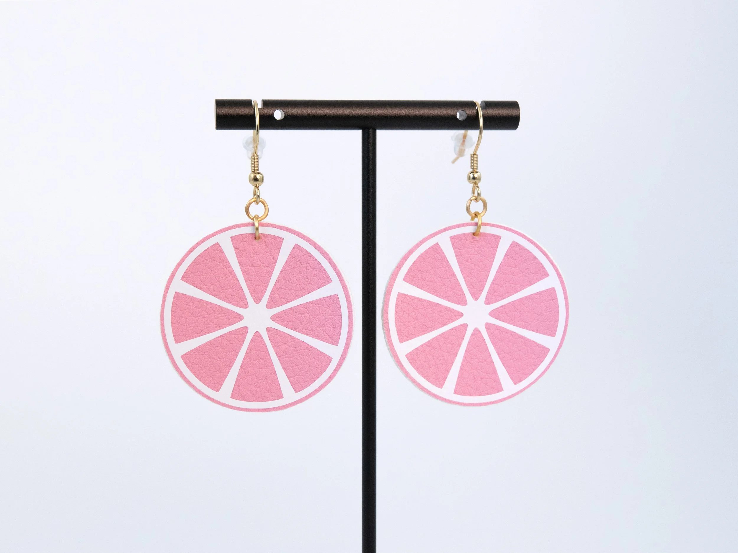 Round citrus slice earrings on a T-bar display stand. The earrings have pink faux leather, white vinyl details, and gold-finished hooks