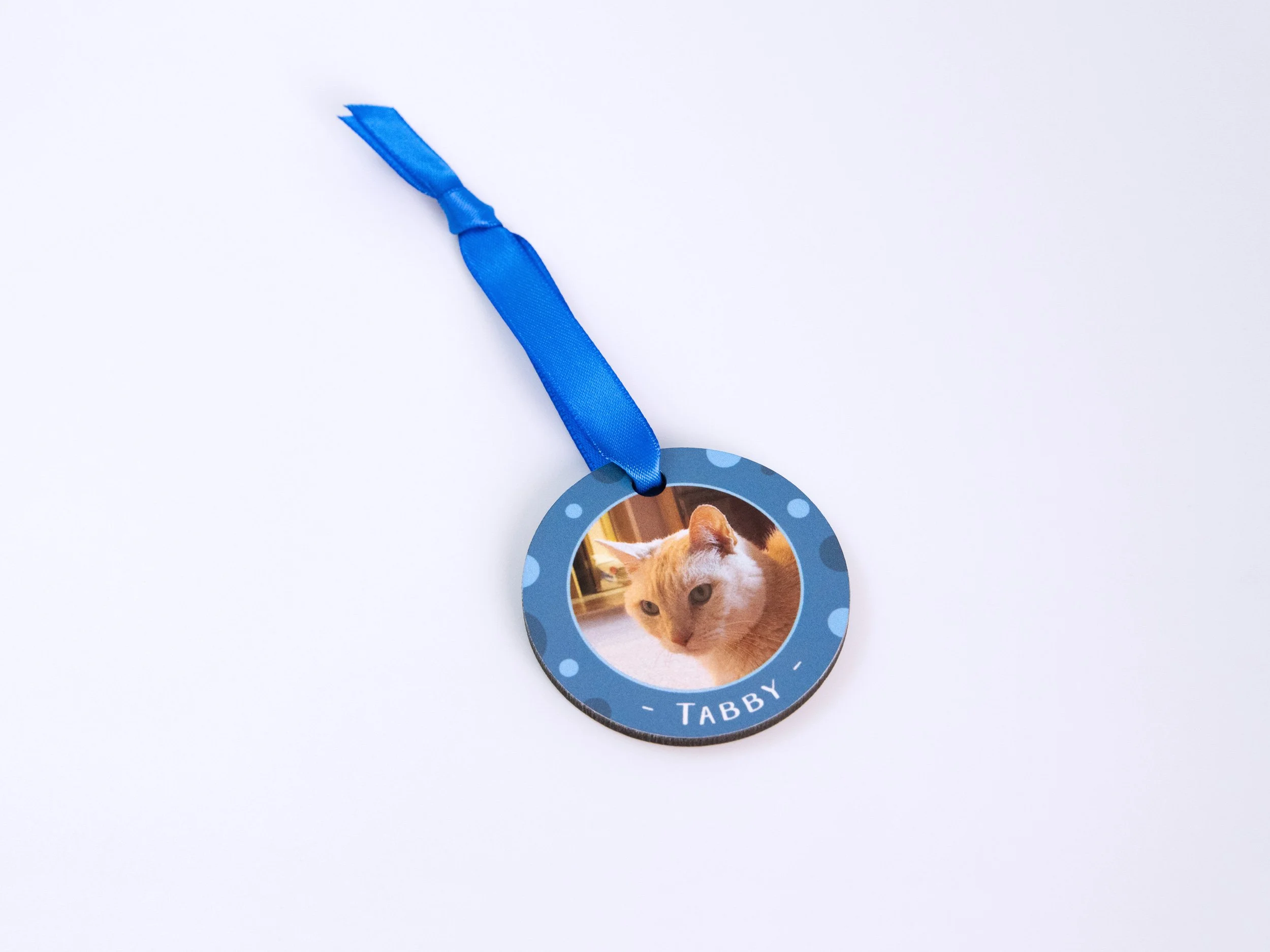 Top view of a sublimated MDF ornament with a ribbon. It has a tan striped cat photo, blue color scheme, and the name "Tabby"