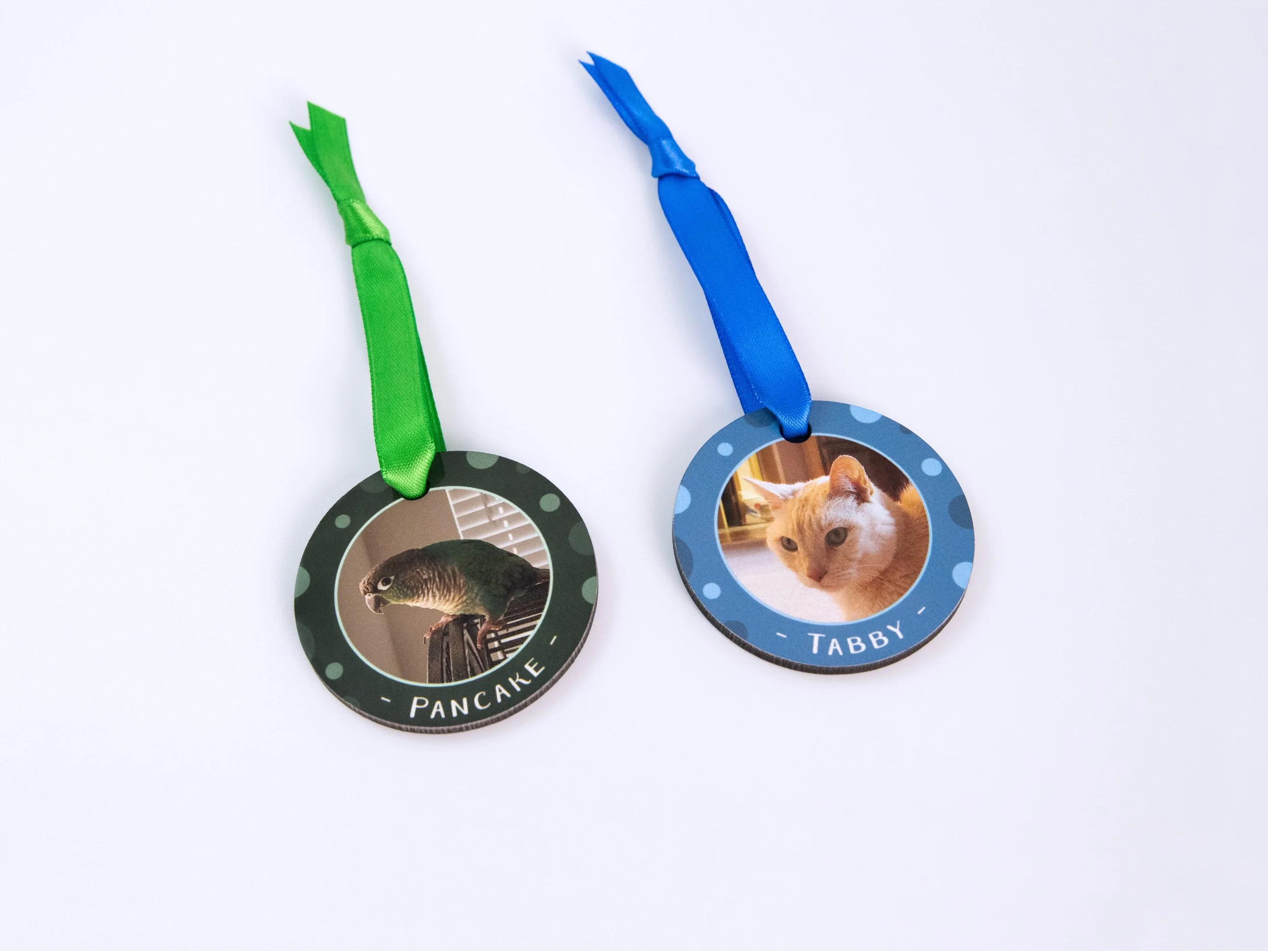 Top view of two sublimated MDF ornaments with ribbons. The left design has a conure photo, green color scheme, and the name "Pancake." The right design has a tan striped cat photo, blue color scheme, and the name "Tabby"