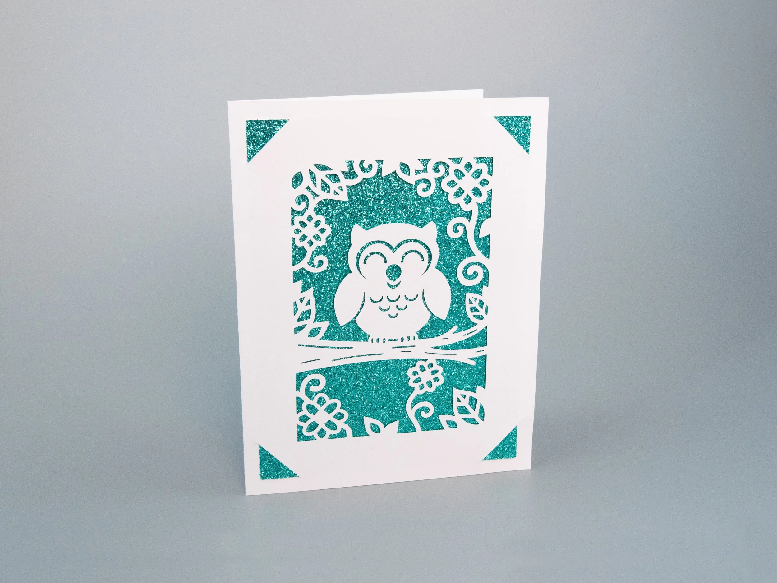 Angled view of a greeting card with an owl cutout and turquoise glitter background
