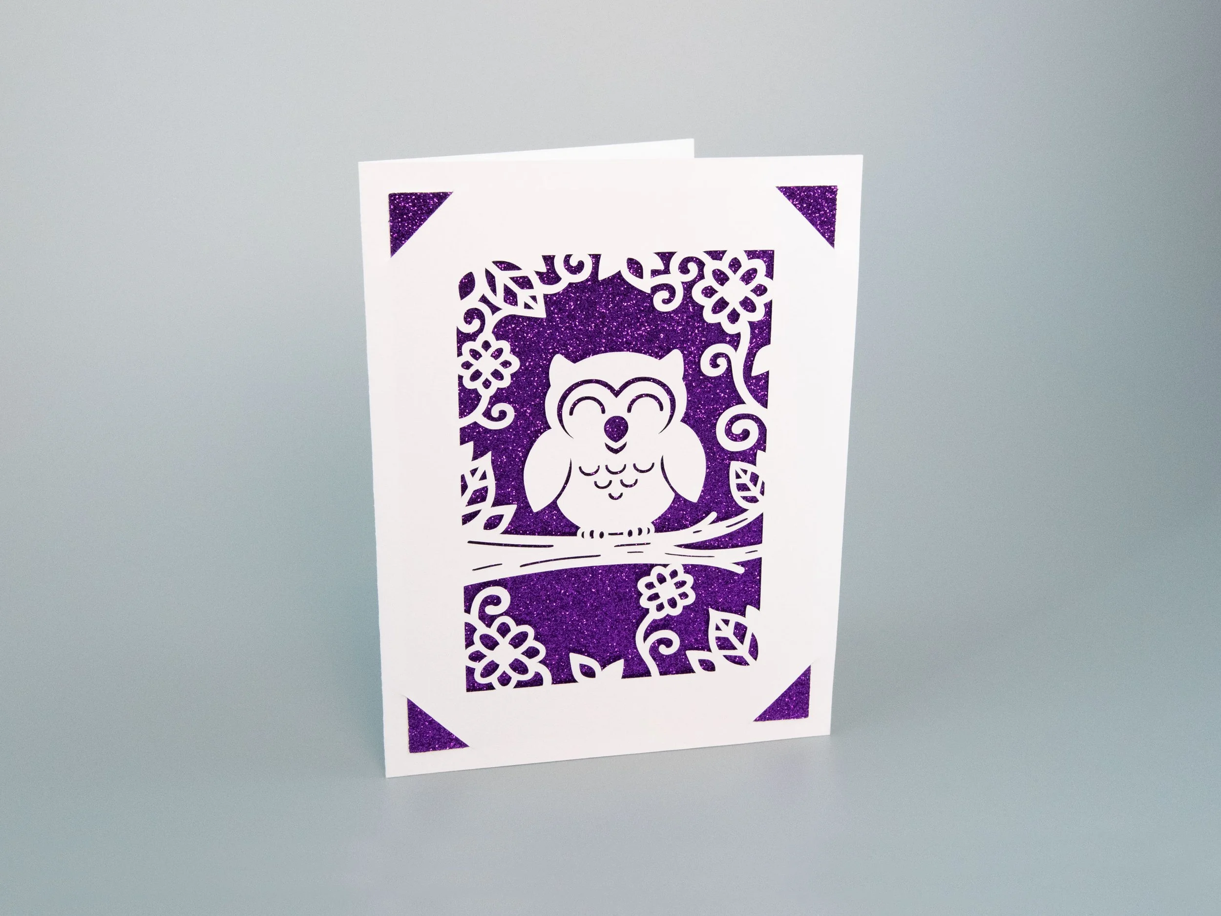 Angled view of a greeting card with an owl cutout and purple glitter background