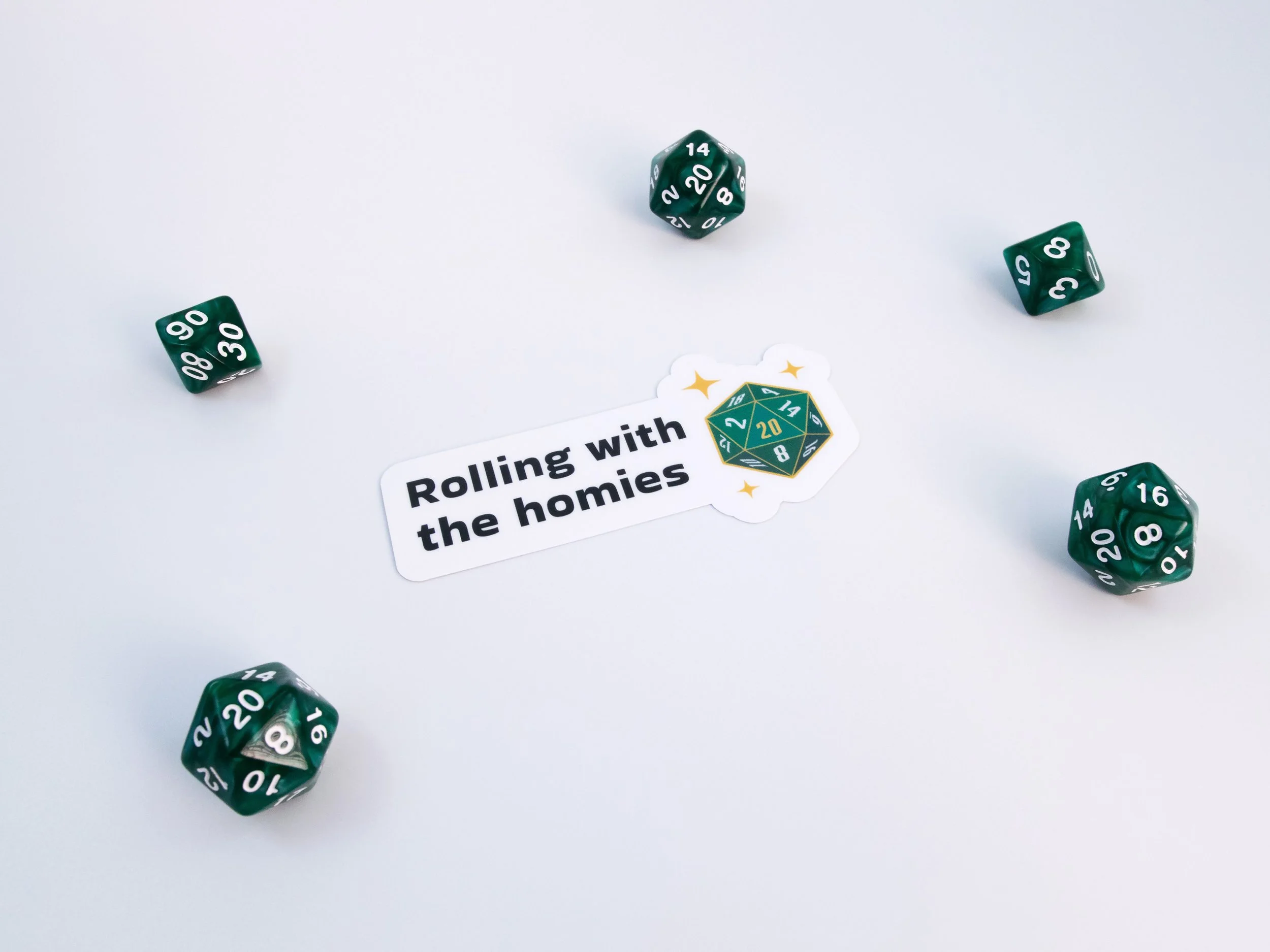 A sticker of a green d20 die with text that says “Rolling with the homies”. The sticker is surrounded by green dice