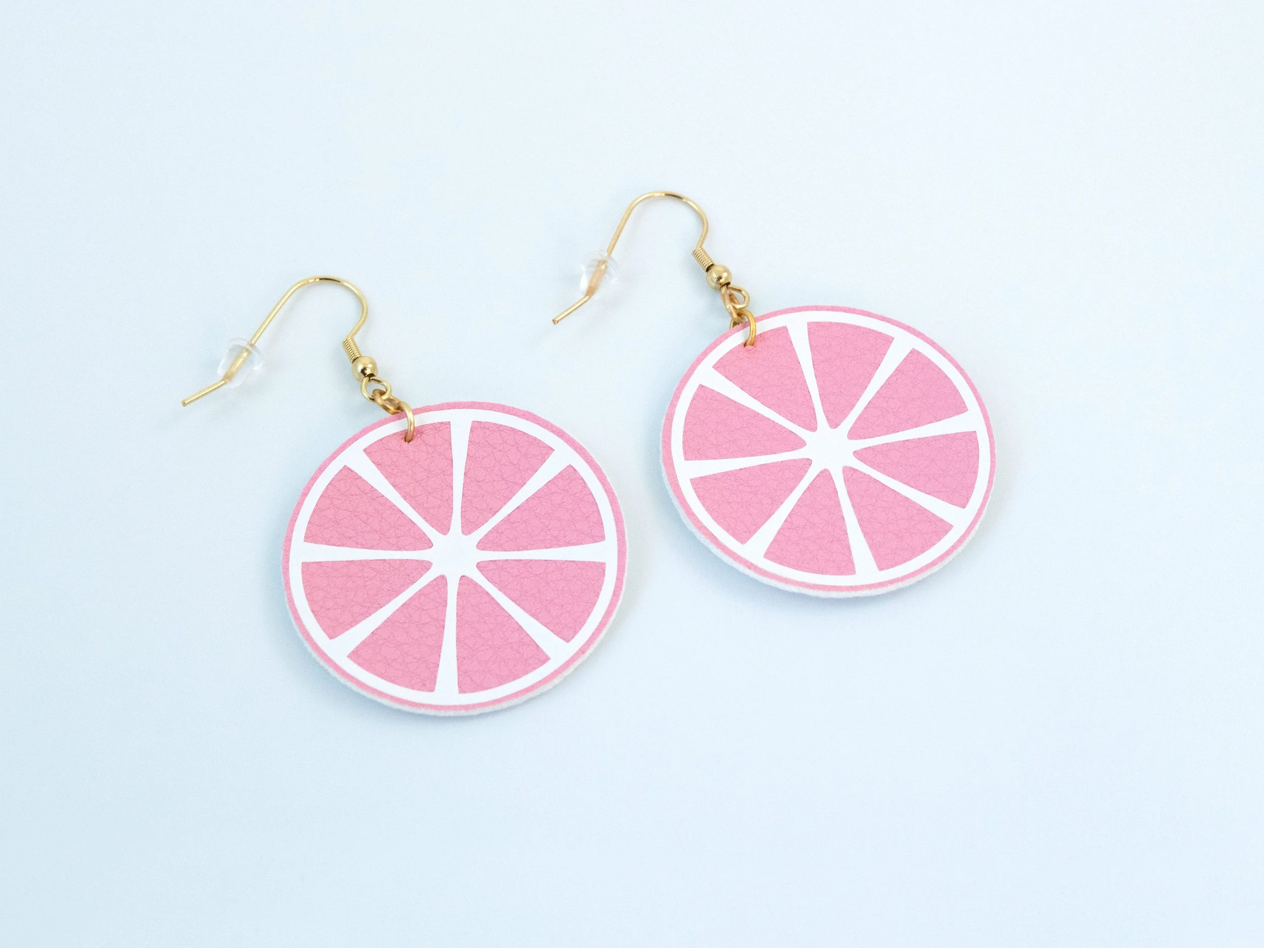 Angled view of round citrus slice earrings. They have pink faux leather, white vinyl details, and gold-finished hooks