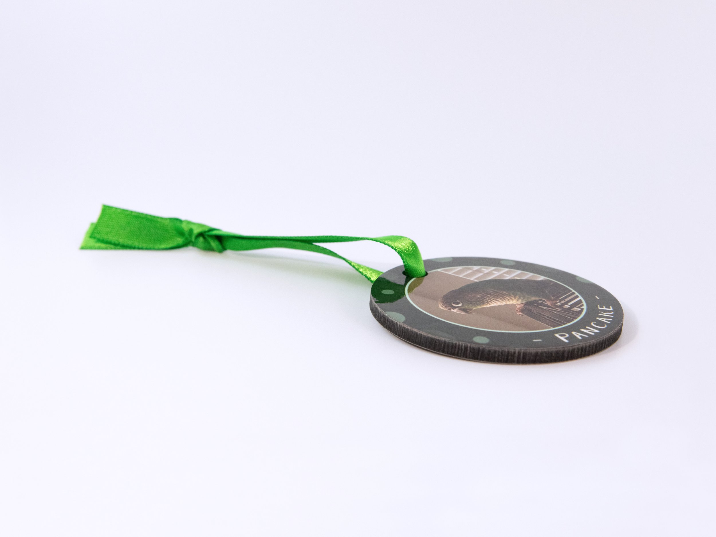 Close up side view of a sublimated MDF ornament with a ribbon. It has a conure photo, green color scheme, and the name “Pancake”