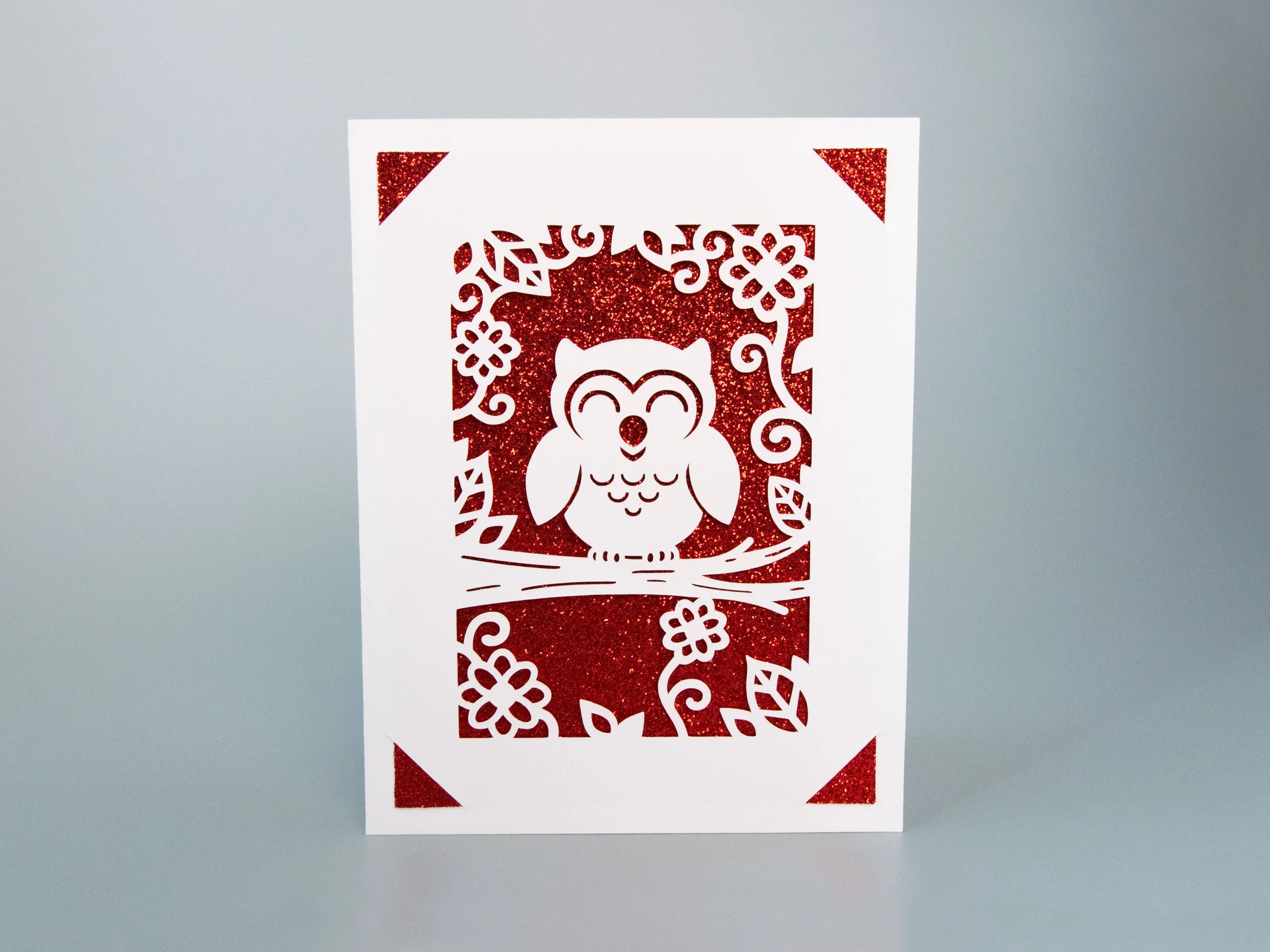 Front view of a greeting card with an owl cutout and red glitter background