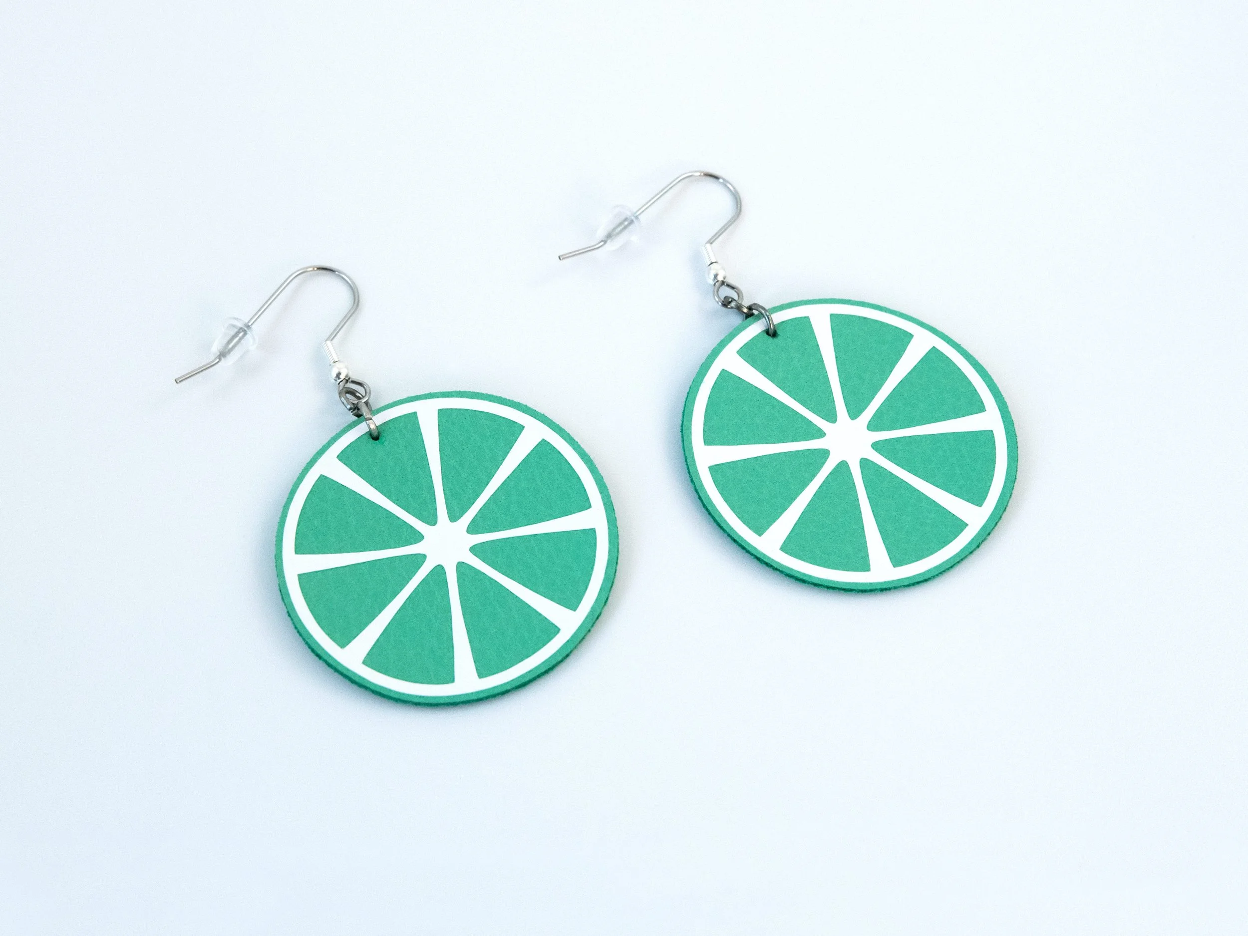Angled view of round citrus slice earrings. They have green faux leather, white vinyl details, and silver-finished hooks