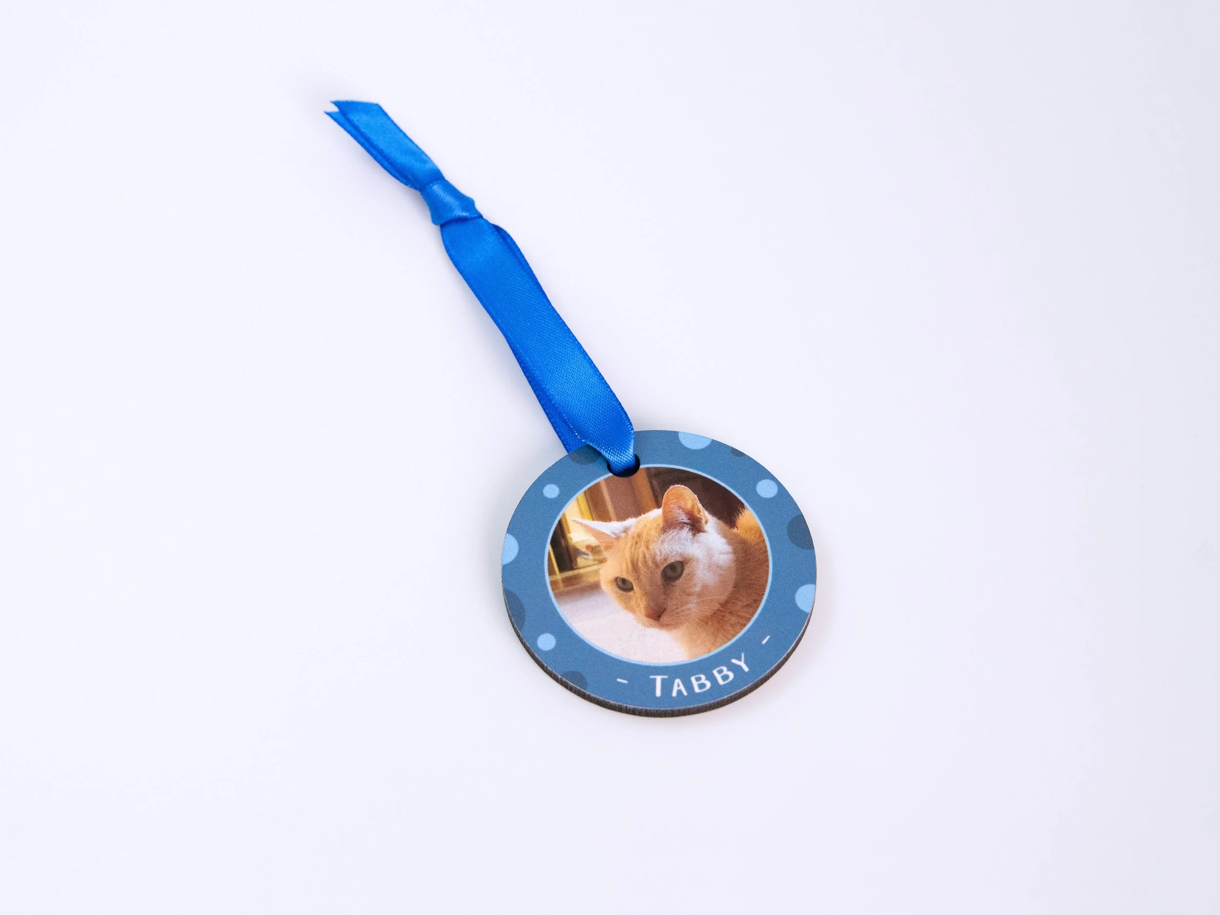 Top view of a sublimated MDF ornament with a ribbon. It has a tan striped cat photo, blue color scheme, and the name “Tabby”