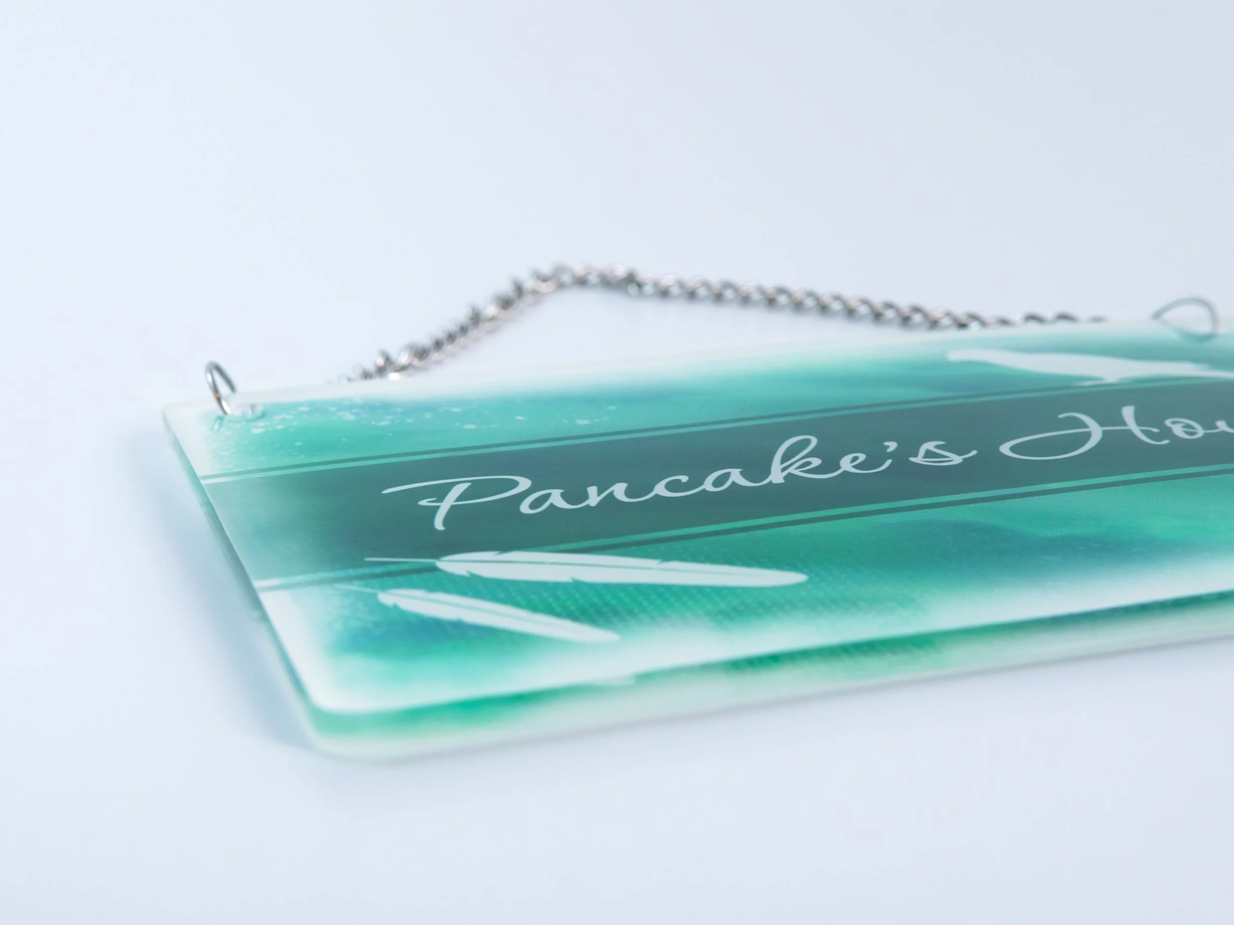 Close up corner view of a small acrylic sign with a metal chain. It has bird-themed artwork with text that says "Pancake’s House"