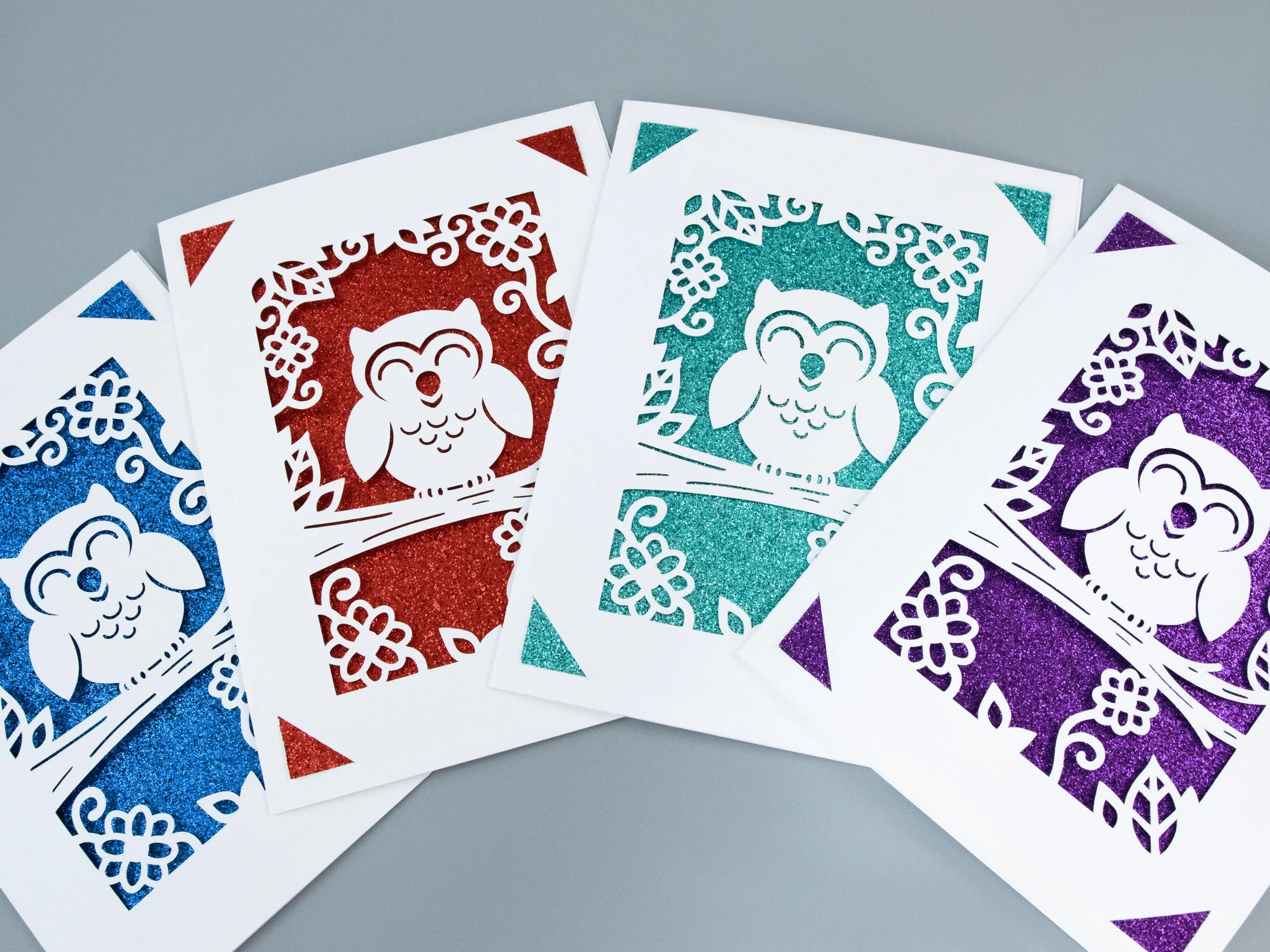 Top view of 4 owl-themed greeting cards fanned out across a table. Each card has a different glitter background color